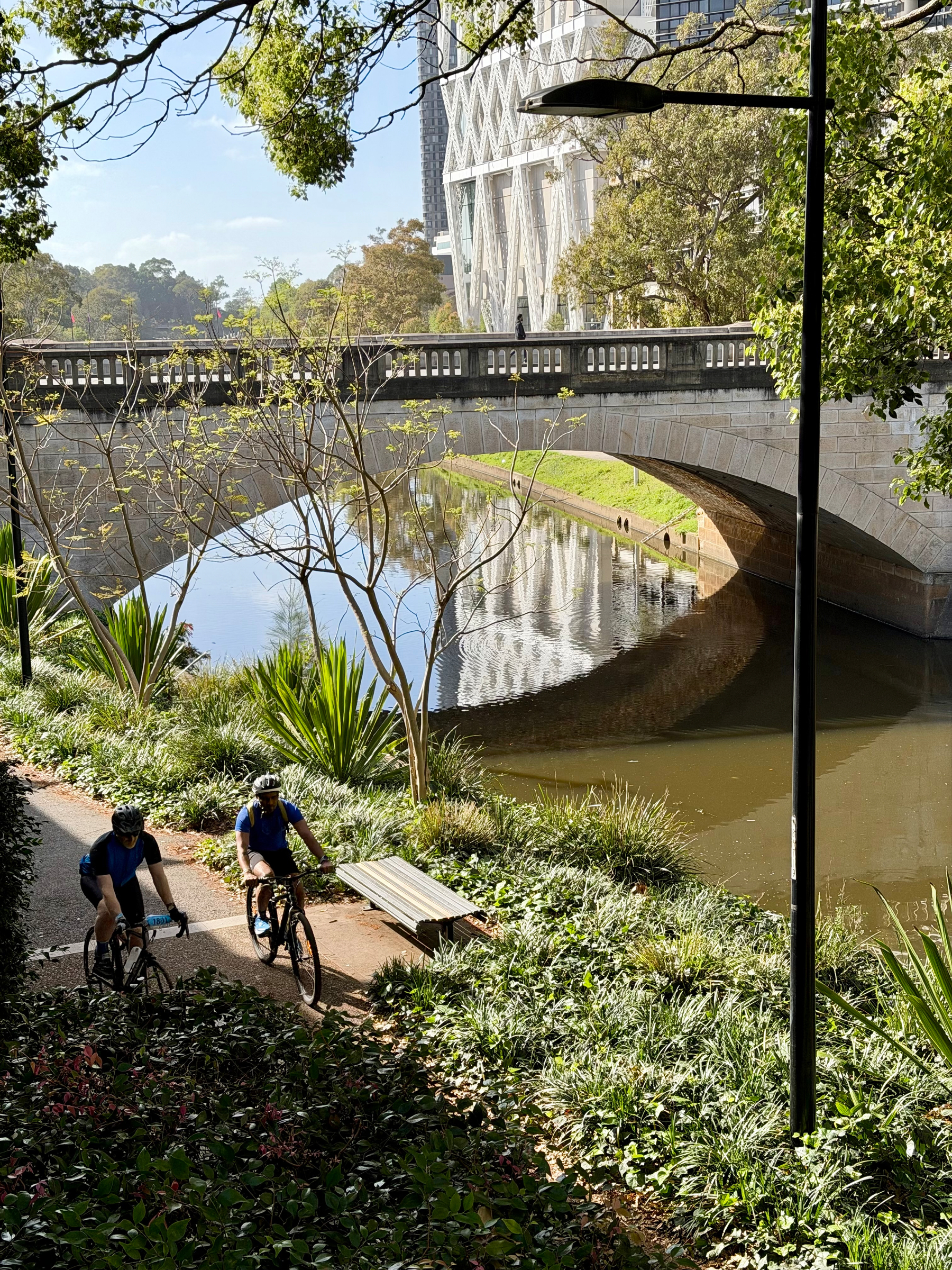 Two cyclists ride along a shaded path beside a river, near a stone bridge and surrounded by greenery. It’s the Parramatta River at Parramatta. 