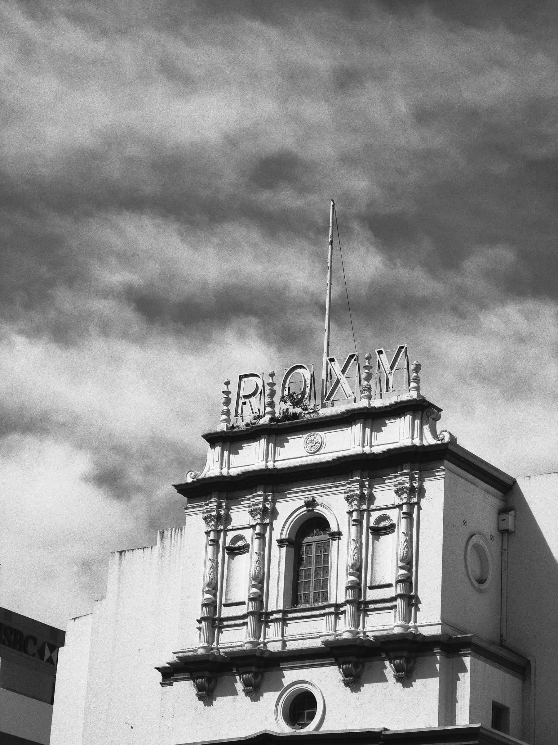 A historic building with classical architectural details features a rooftop sign spelling out ROXY against a cloudy sky. Black and white photo of the top part of the Roxy Theatre in Parramatta.  