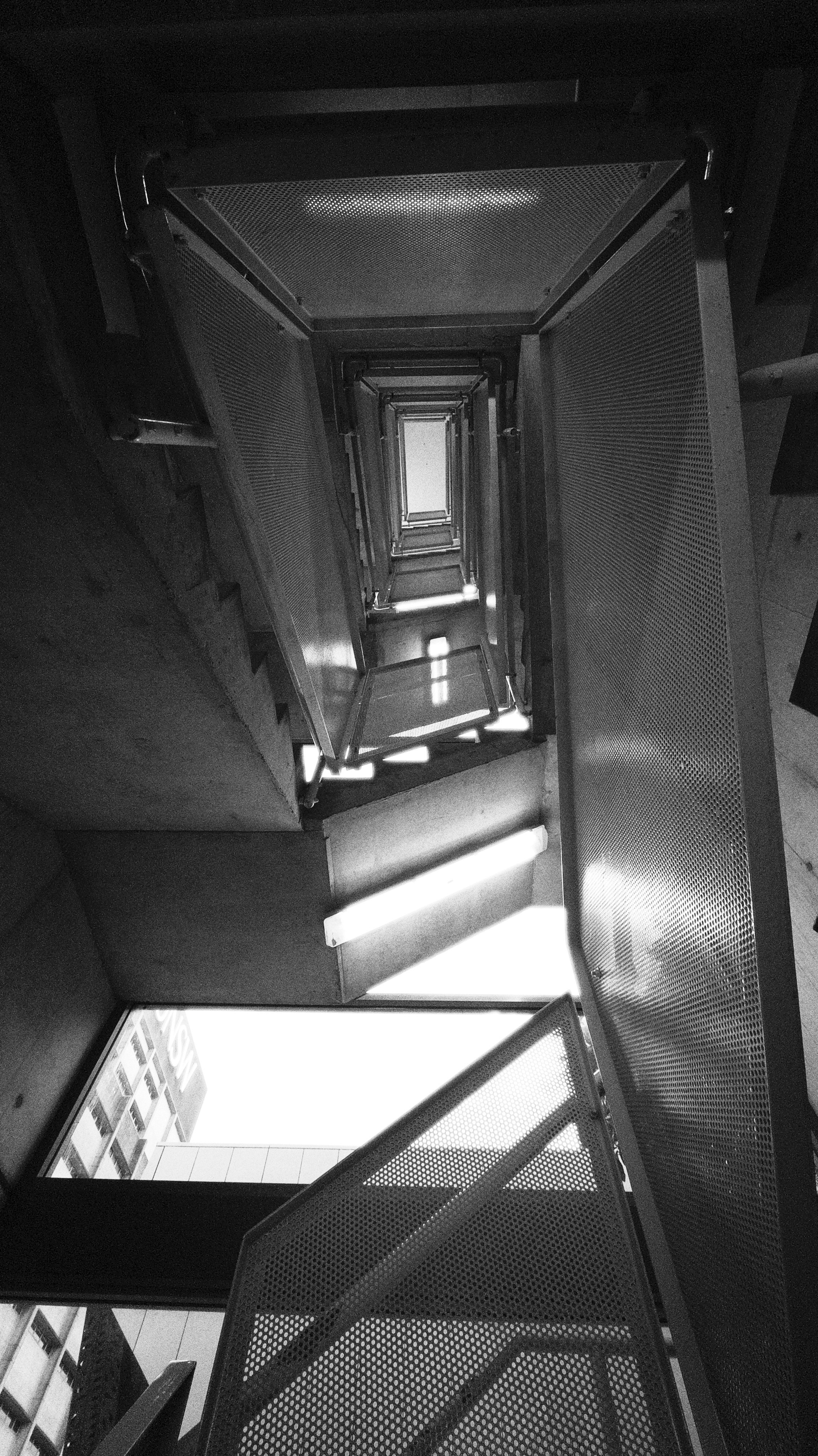 A black and white photo captures a stairwell from the bottom, looking upward through metal mesh railings with light from above.
