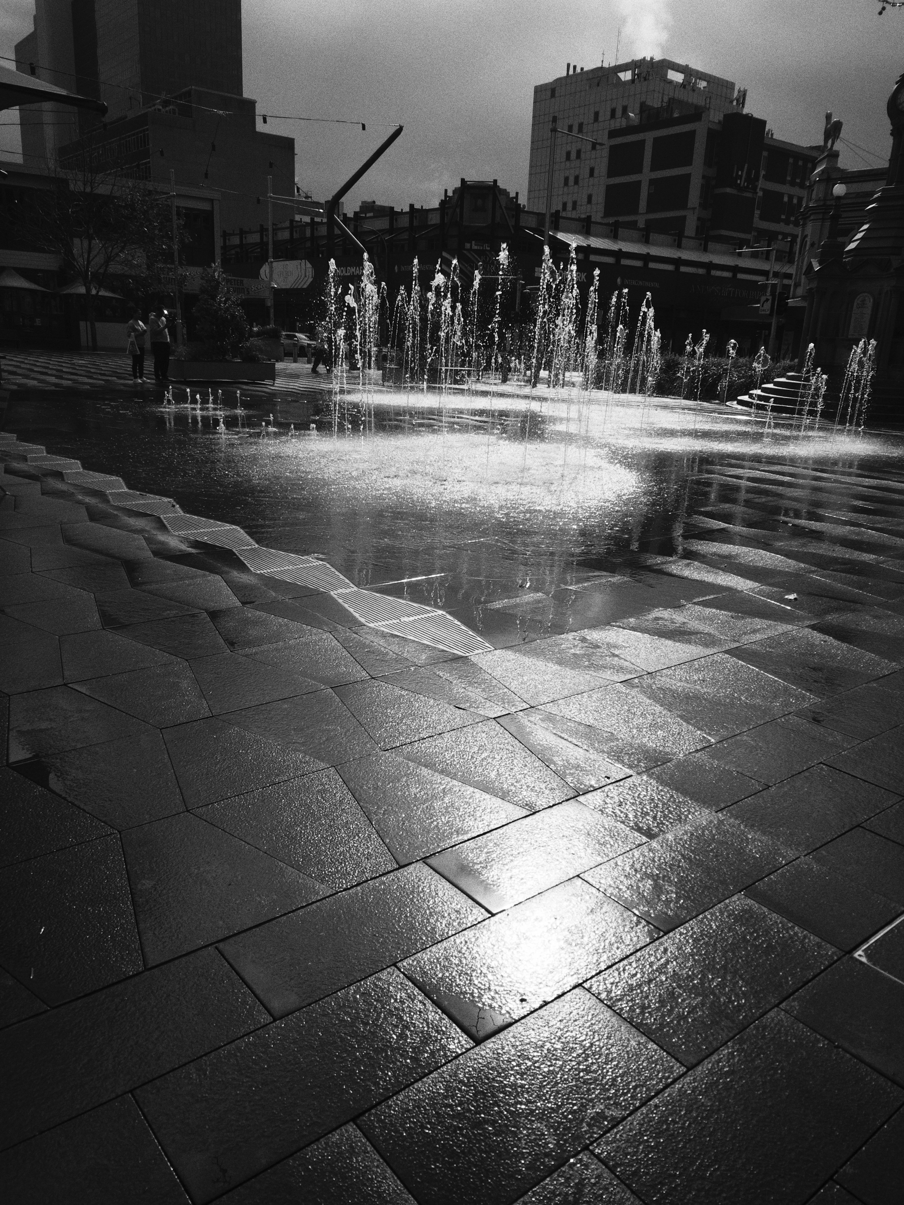 A series of water fountains spray upward in an urban plaza with wet, reflective stone pavement and tall buildings in the background.