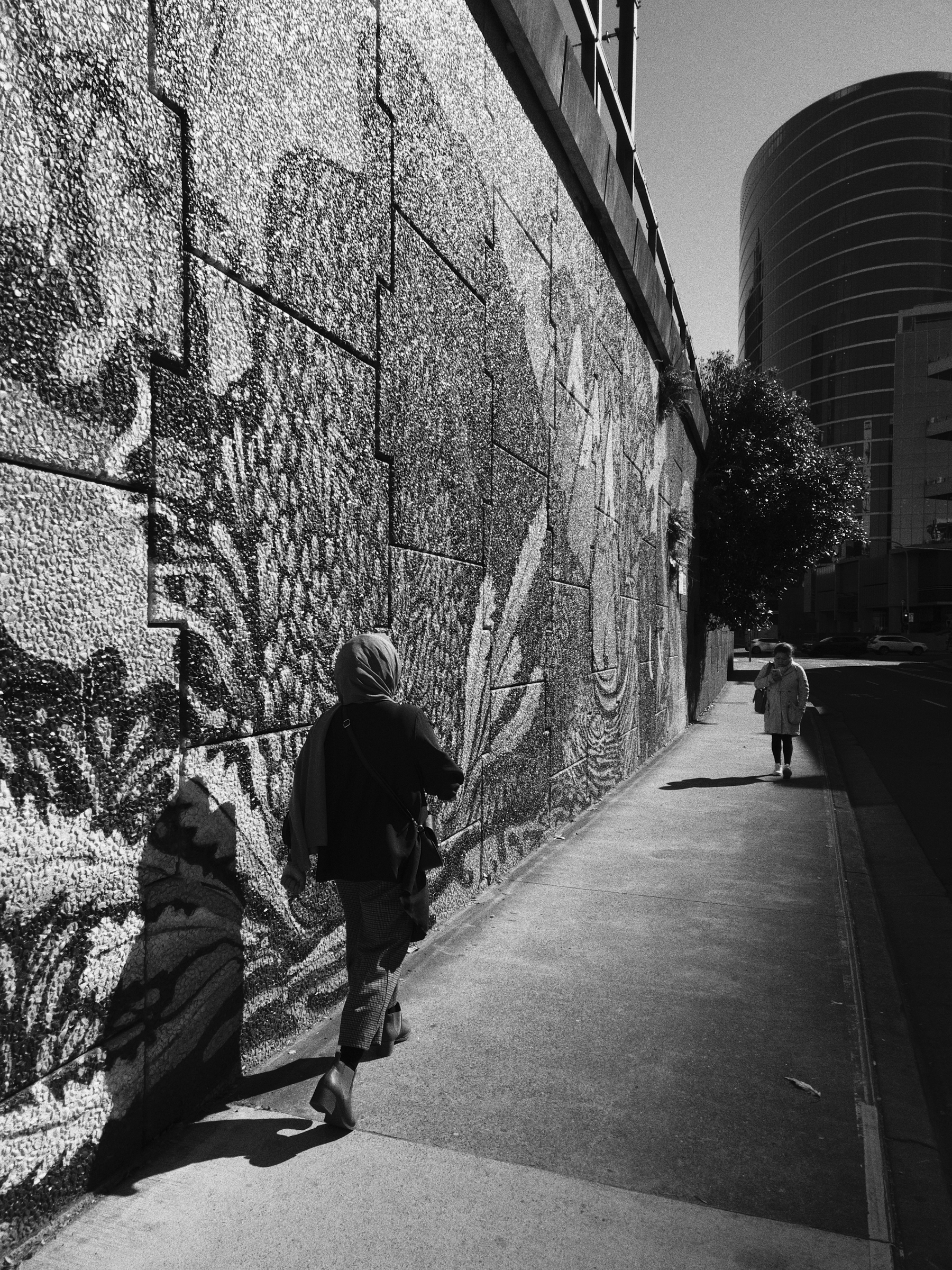 Two people are walking along a sidewalk next to a large textured mural wall in an urban environment under bright sunlight.