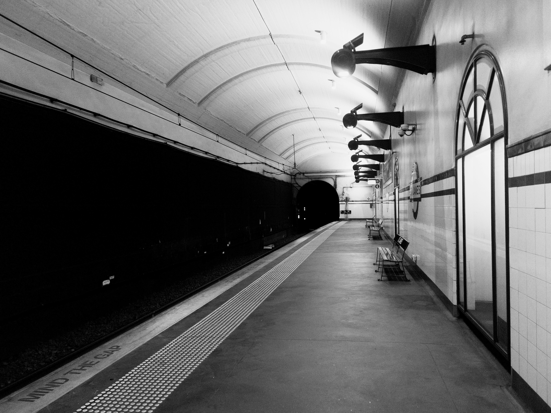 A deserted underground train station platform is illuminated by wall-mounted lamps, featuring a curved ceiling and a dark tunnel at the far end.