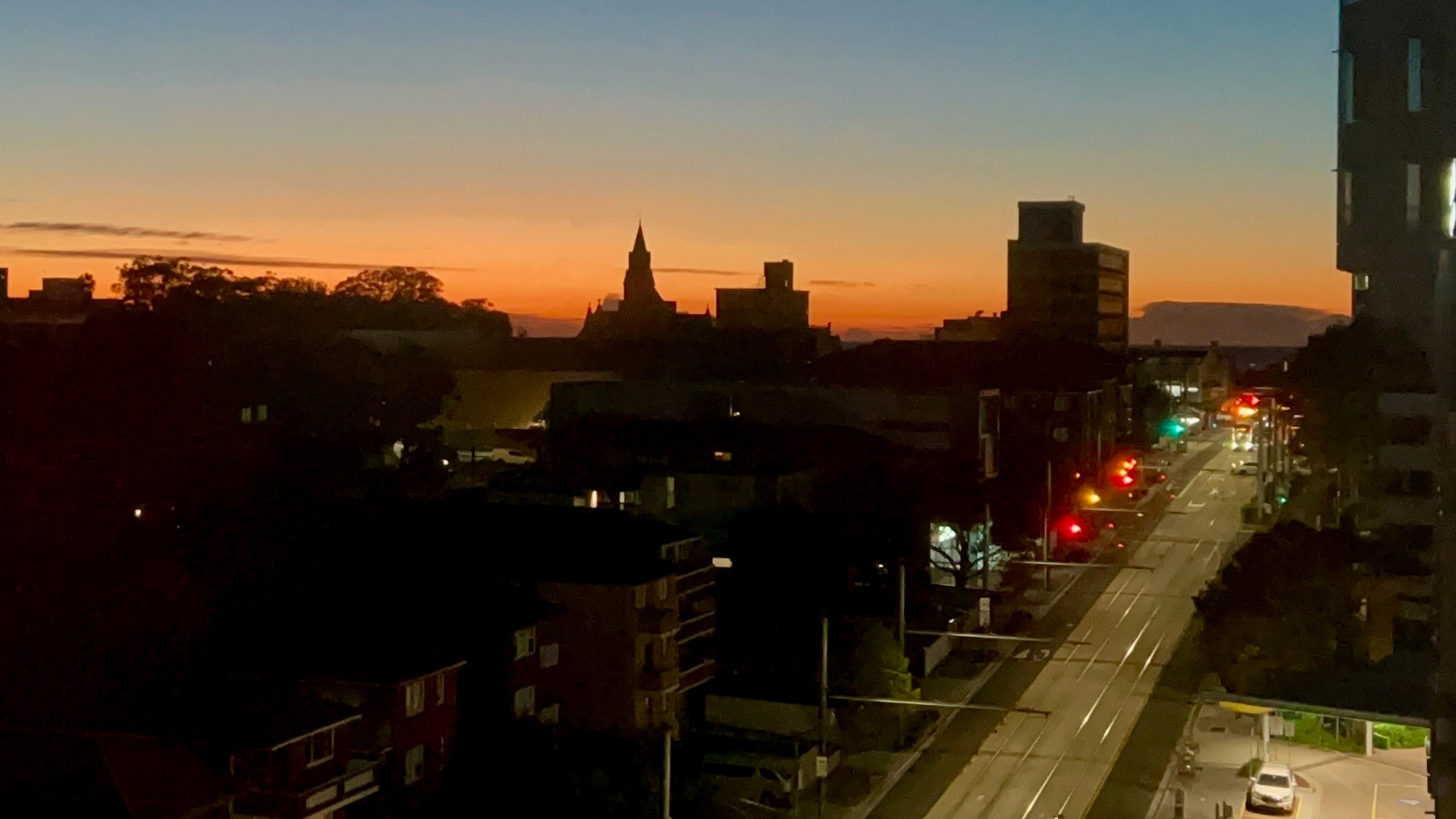 City street with tram tracks, glowing traffic lights, and silhouetted buildings against an orange dawn sky