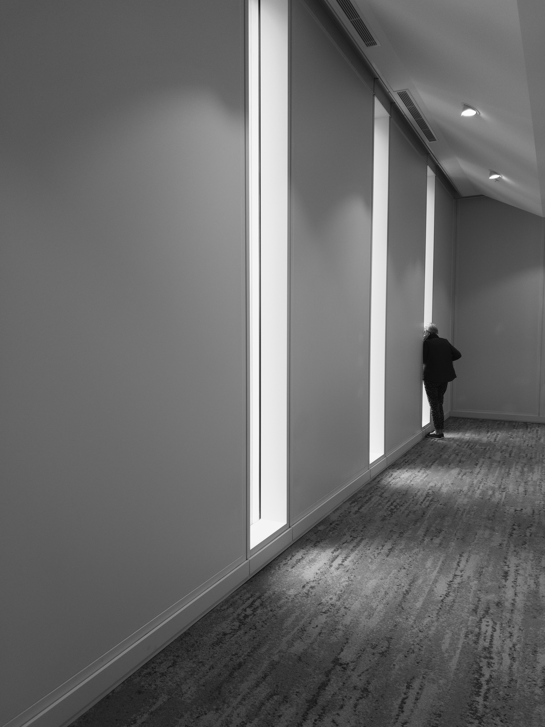 A solitary person stands by tall, narrow windows in a dimly lit, minimalist hallway.