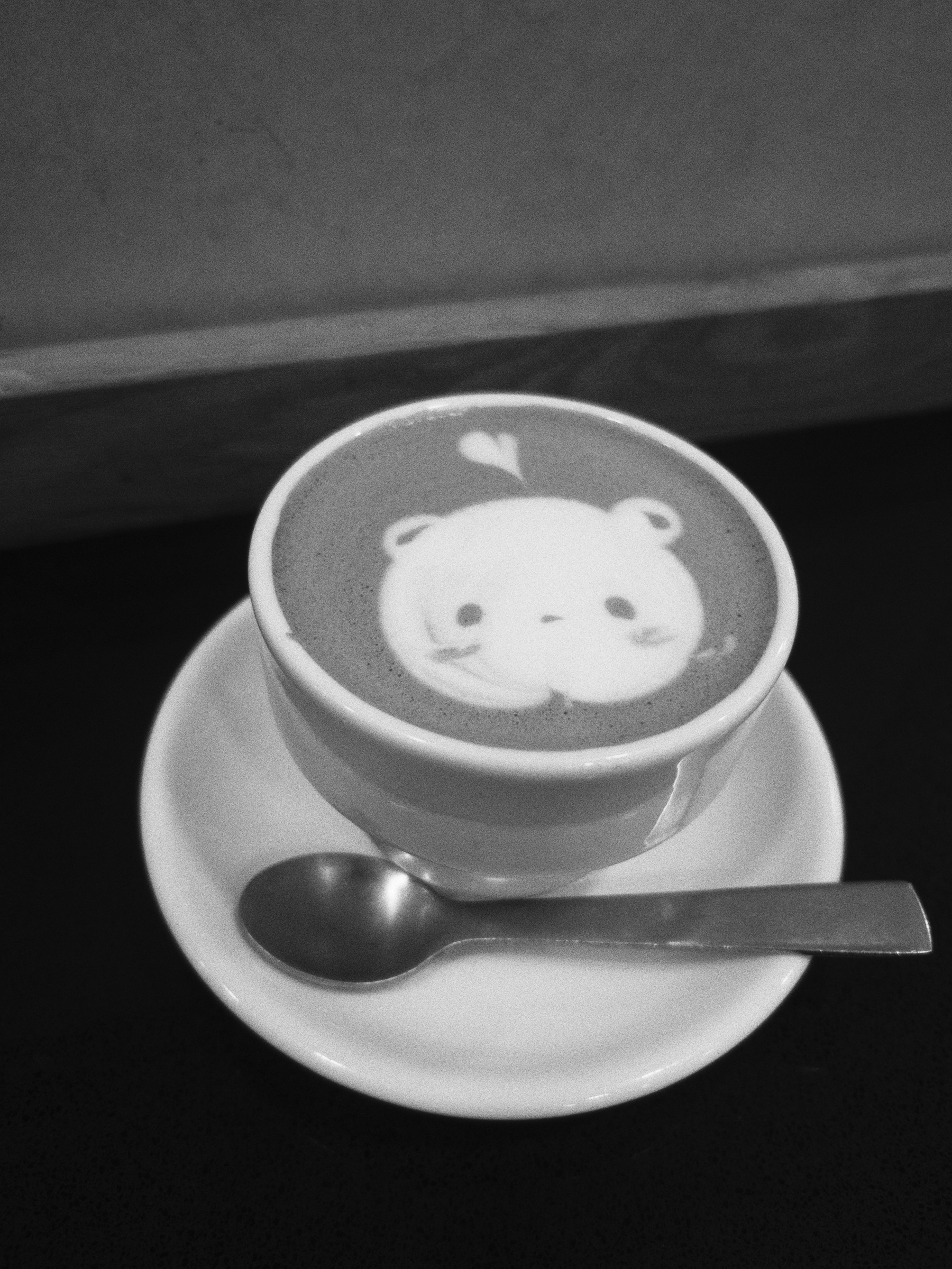 A cup of coffee features latte art depicting a cute bear face with a small heart above it, accompanied by a spoon on a white saucer.