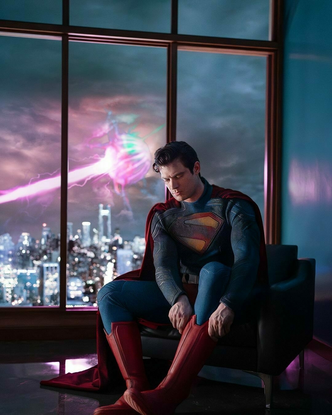 The first look at James Gunn's Superman, as we see David Corenswet, in costume, sitting in a chair, pulling on his boots, as some sort of laser explosion occurrs in the window behind him.