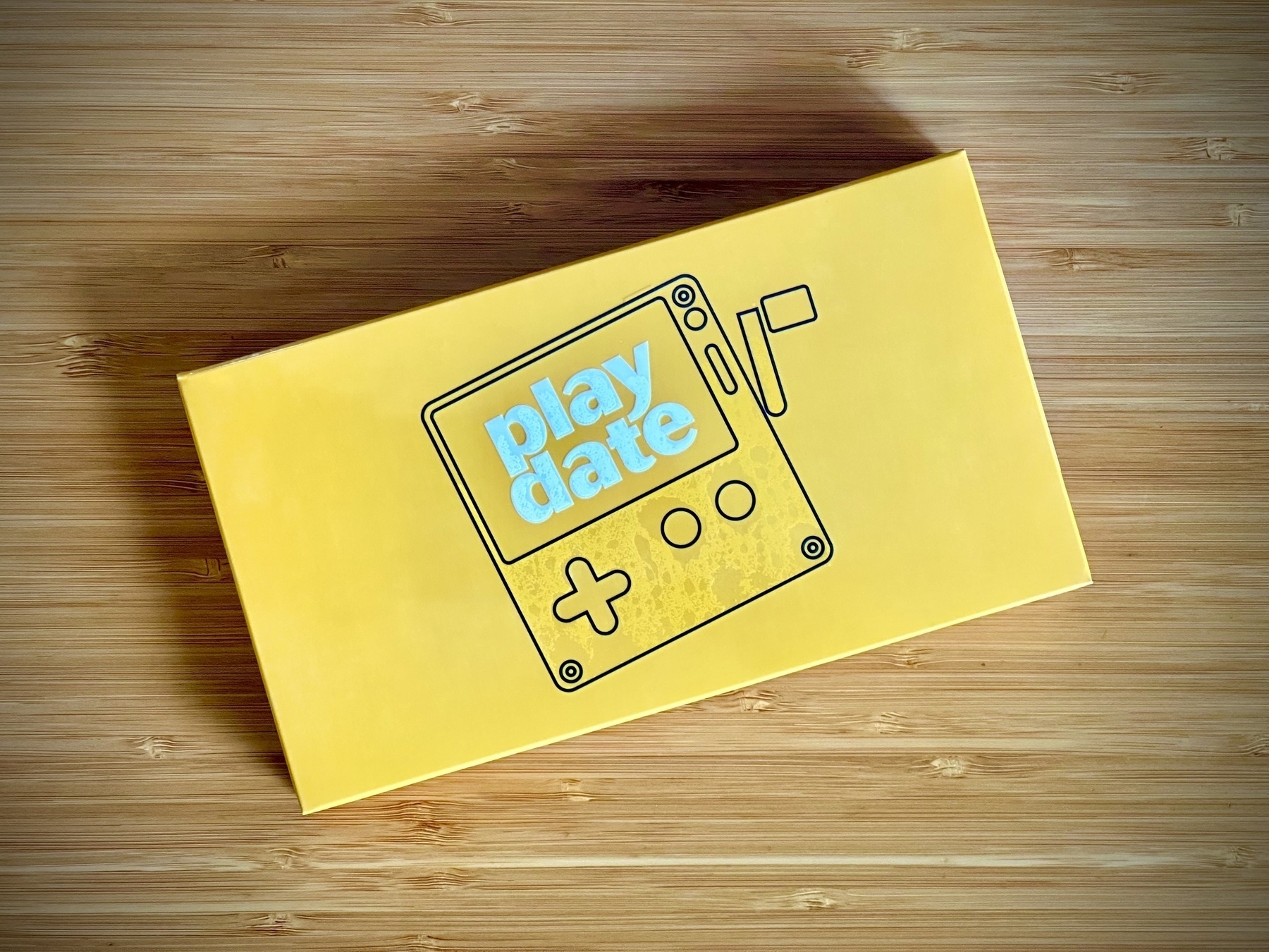 A closeup of a bright yellow boxed Playdate games console on a bamboo desk.