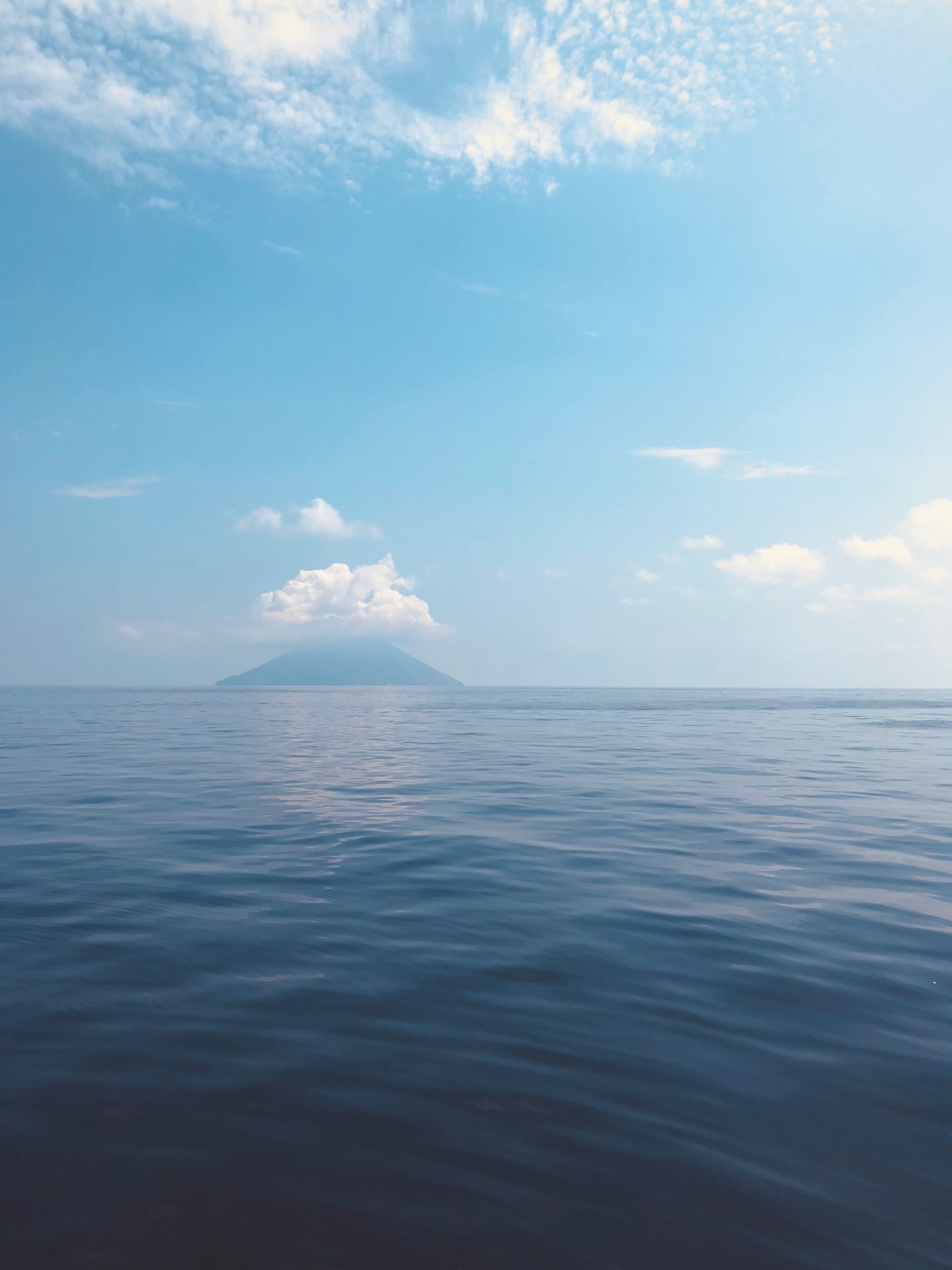 A calm blue ocean stretches to the horizon where clouds obscure the crater of Stromboli.
