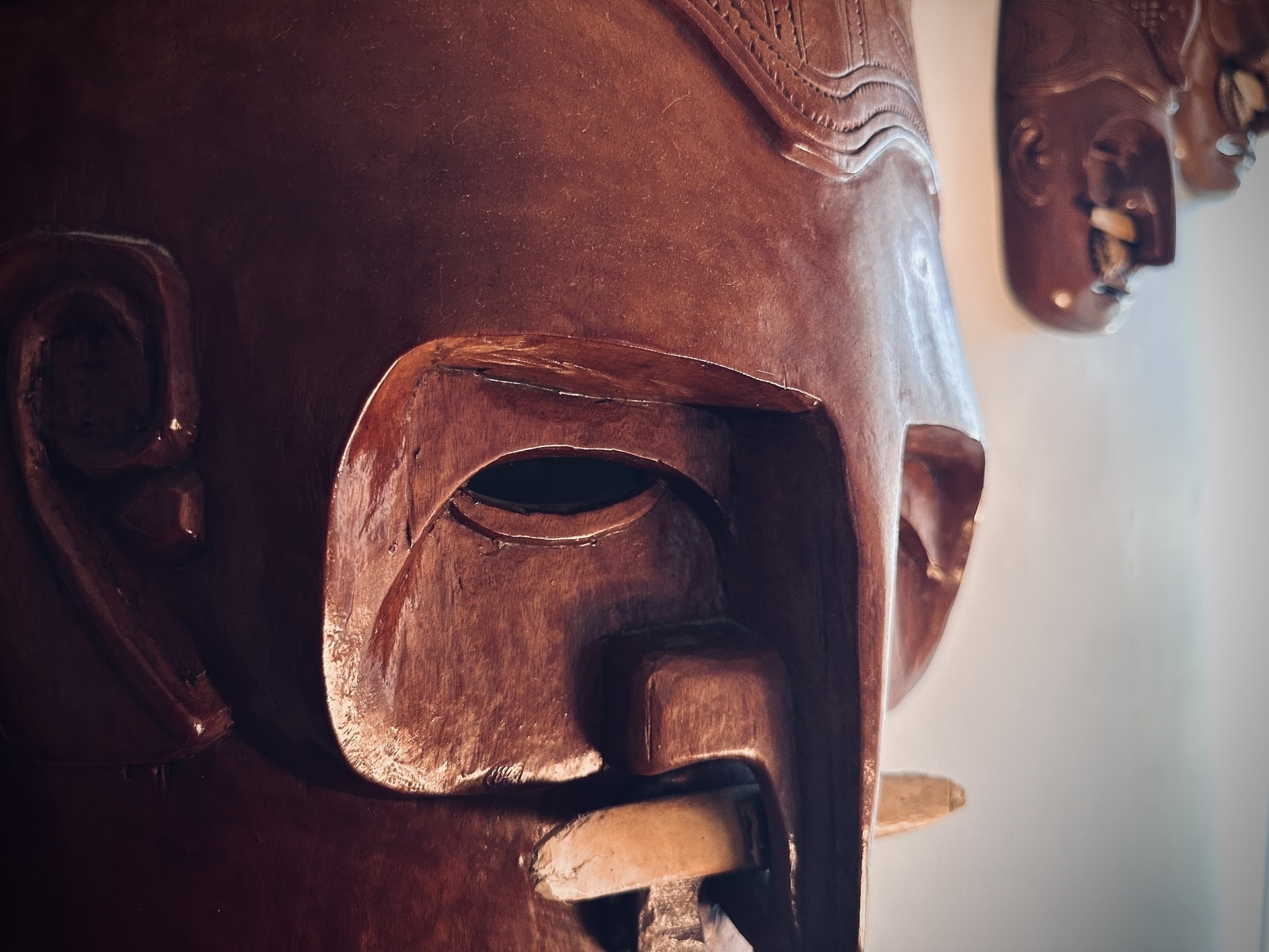 A close up of the carved eye sockets of an old wooden ceremonial mask. A bone cross piece slices through its nose, held firm by carved wooden “shells”.&10;&10;Smaller companions hang behind it, staring. 
