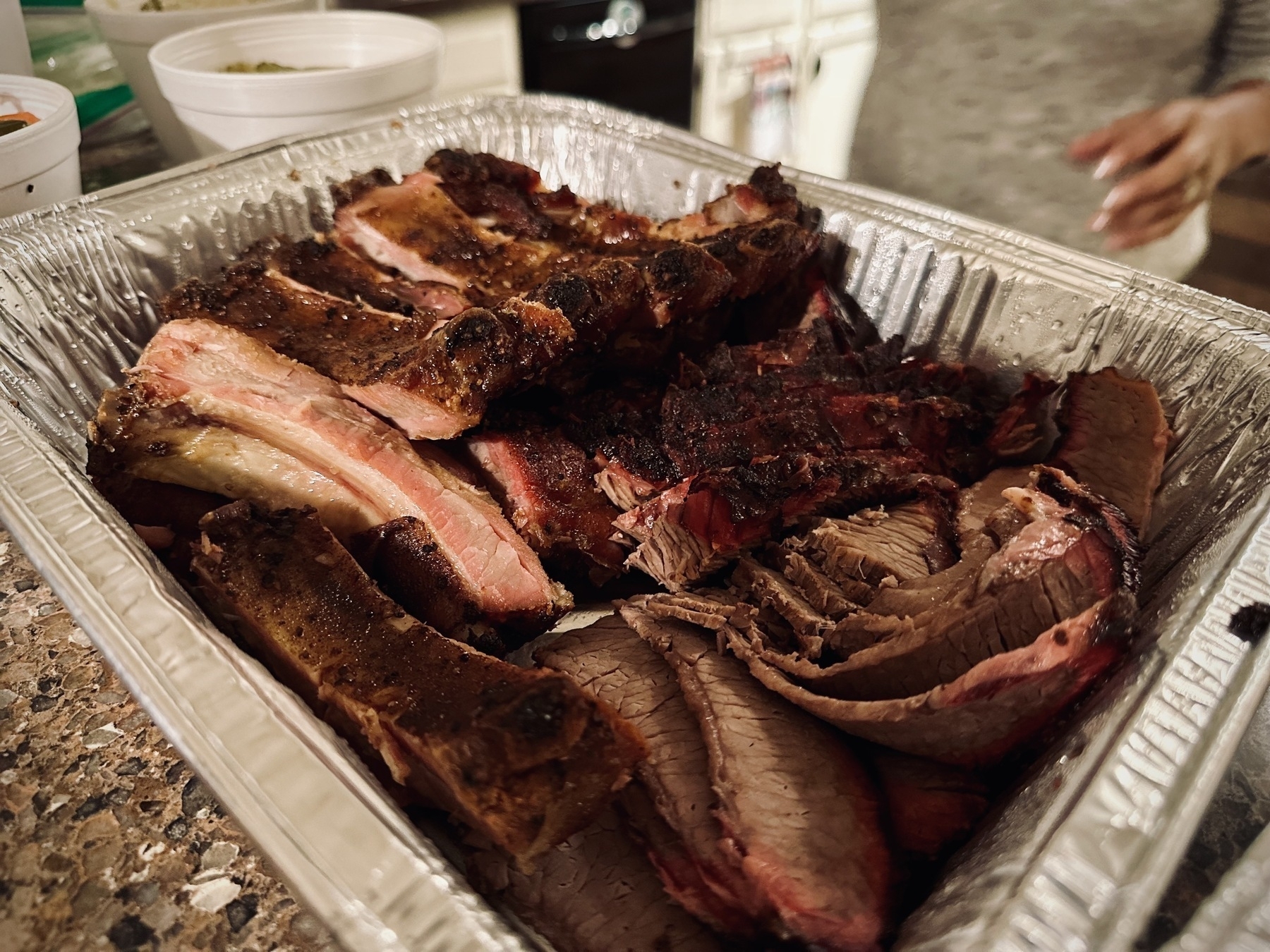 Close up of a box of brisket and ribs from Rudy's in Austin, sitting on a stone bench top, with cups of sies in the background.