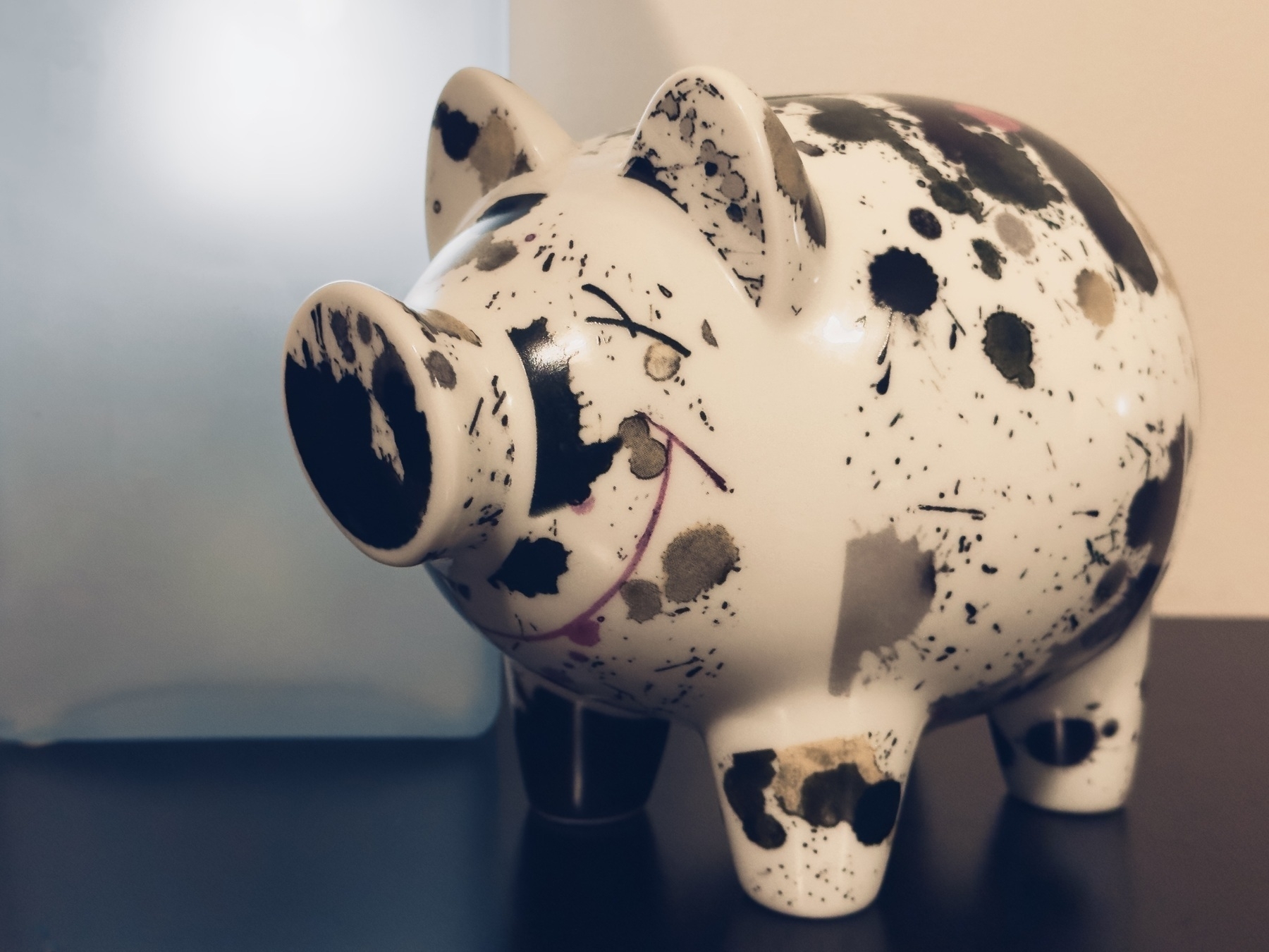 A closeup of glossy white piggy bank decorated with dark paint spatters and smiling as it stands on a dark wooden beside table, backlit by a small lamp.