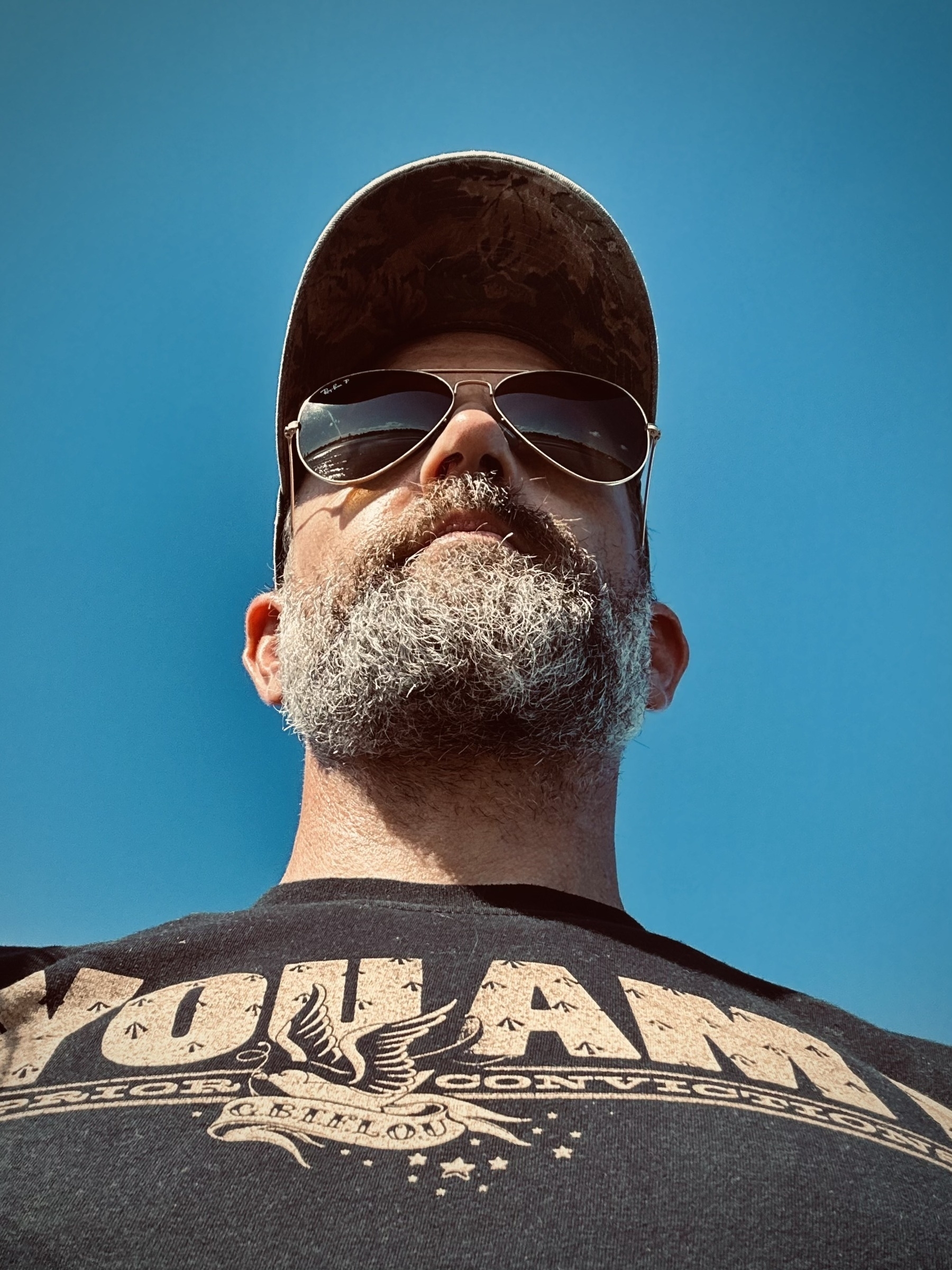 Looking up at the face of 40 something bloke with a greying beard, wearing a You Am I t-shirt and a baseball cap, surrounded by blue sky, an ocean horizon reflected in his aviators.