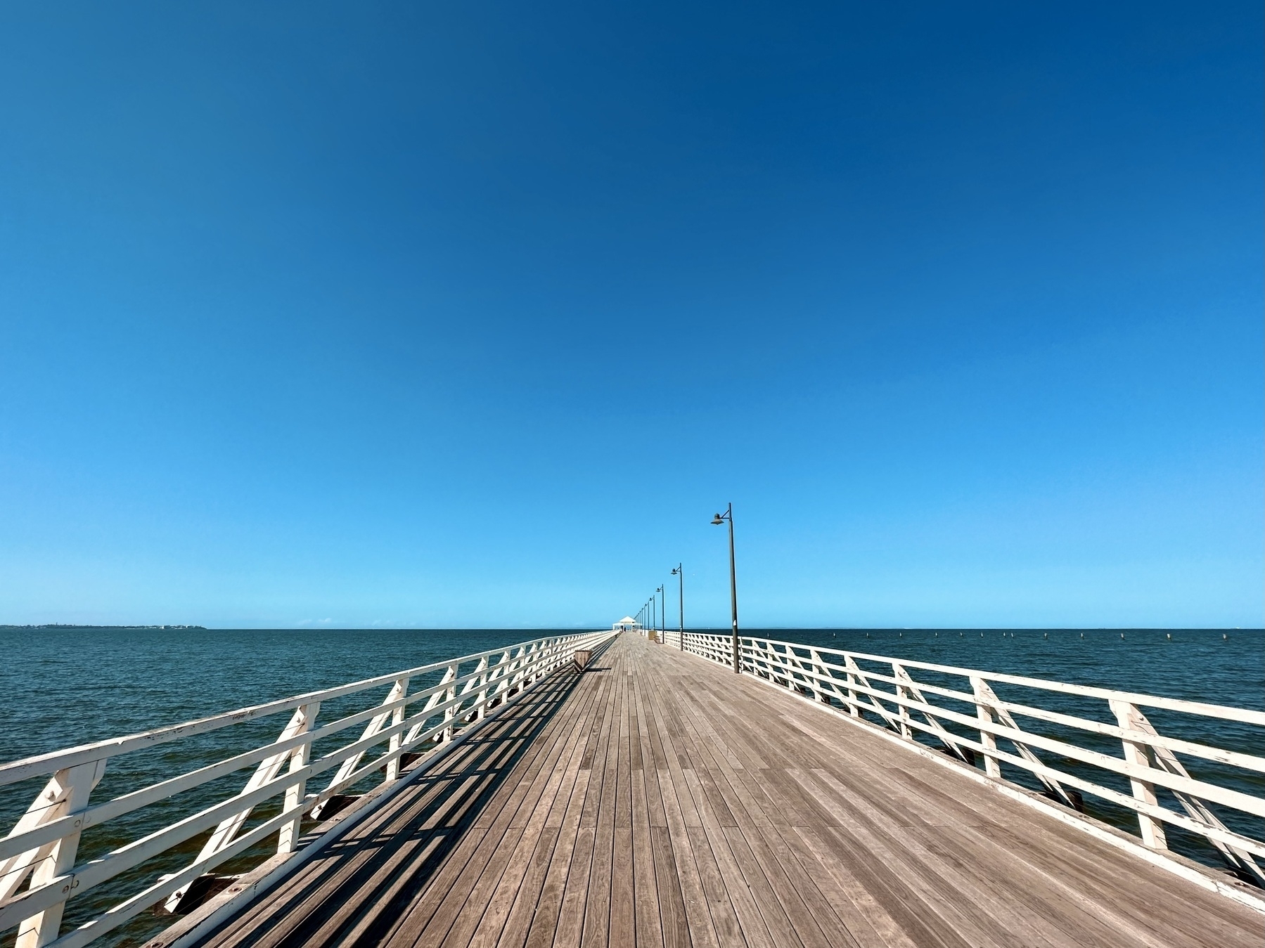 A beautiful clear day, with a bright blue sky. An old wooden pier, with light decking and faded white railings, and lined with old colonial street lamps stretches out toward the horizon, and into the blue green coastal waters. A small gazebo decorating its end sits on the horizon line.