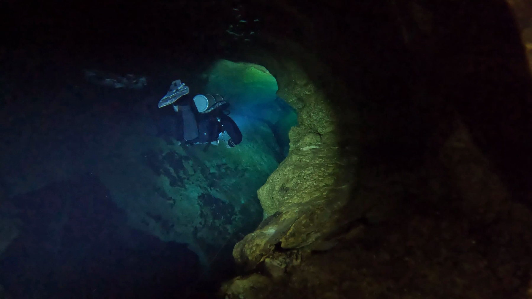 A diver swims down a lava tube in an underwater cave system, his dark drysuit and greyscale camouflage fins lit by a team mates primary light, as it illuminates the walls of the cave in greens and blues.