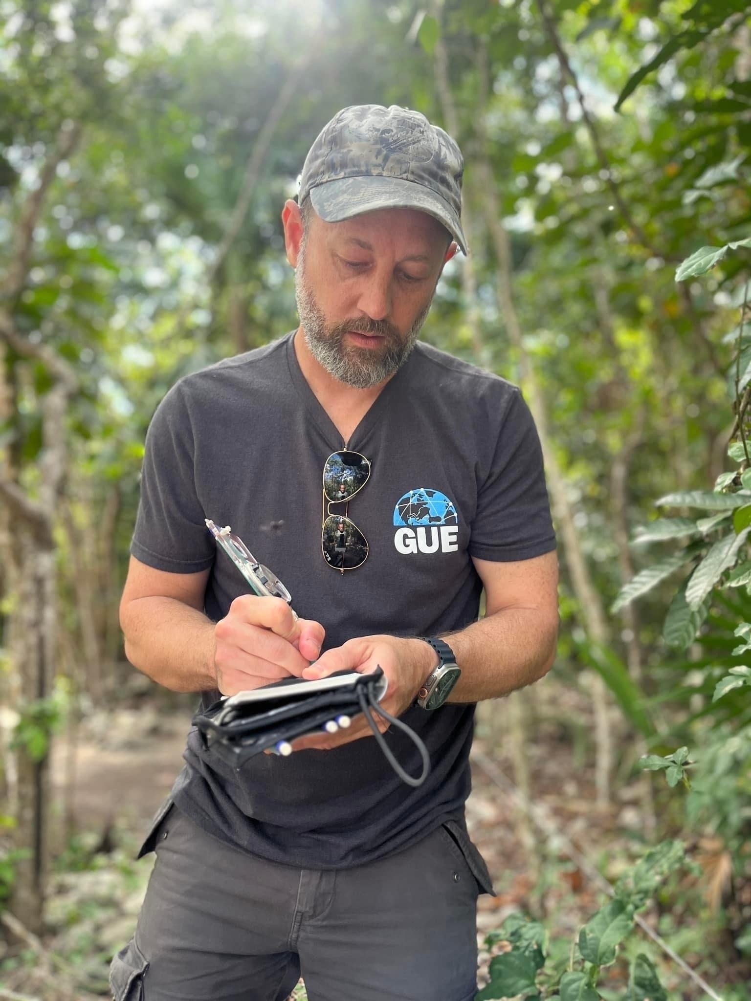 A three quarter shot of a man in a grey t-shirt, cargo pants, and a camouflage hat, standing in the jungle and writing in wetnotes using a pencil with a handheld compass attached to it.
