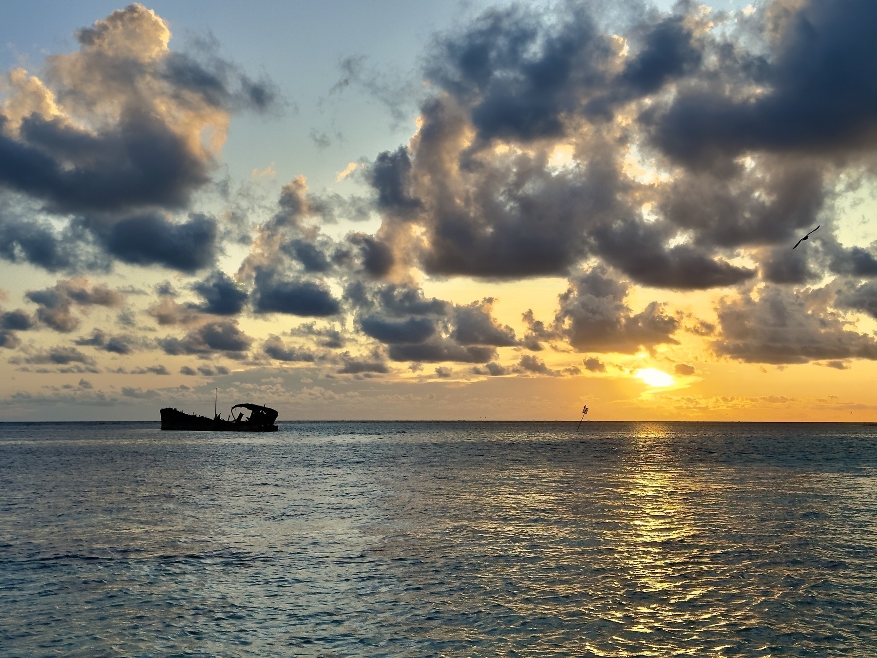 A golden sunset drifts toward an ocean horizon, backlit clouds outlined above a rusted ship wreck jutting above the waterline.
