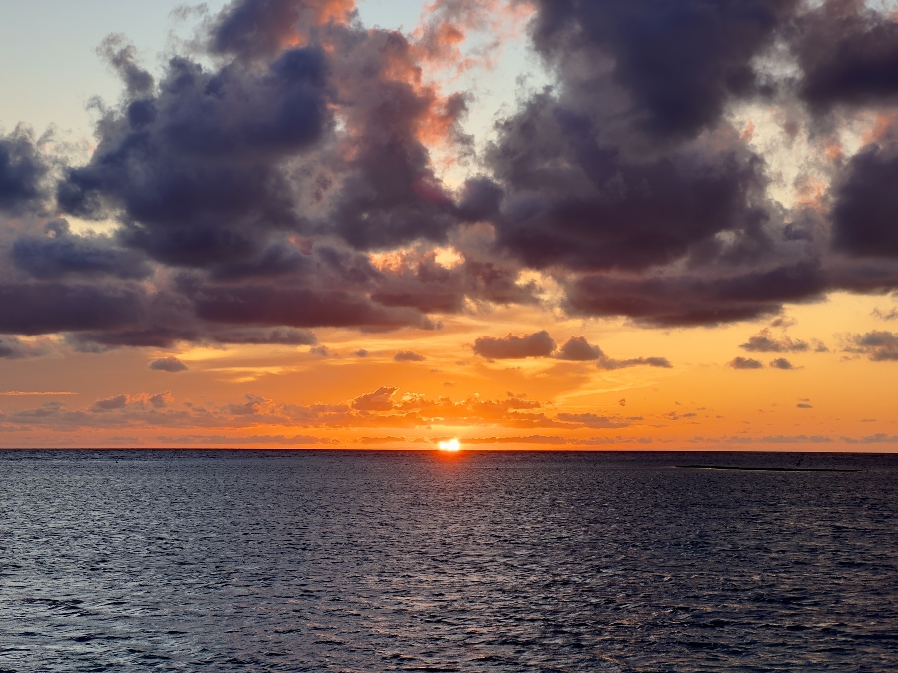 A rich orange sunset, the sun disappearing below an ocean horizon, grey clouds lit with deep purples and oranges.