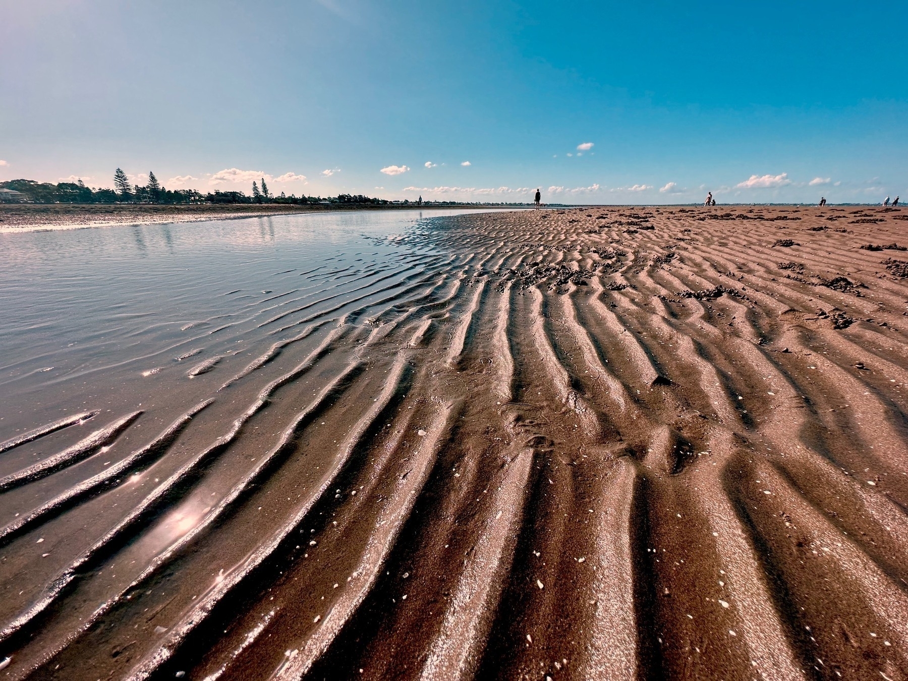 Looking along a beach at low tide, a bright blue sky above wavy patterns in the sand.