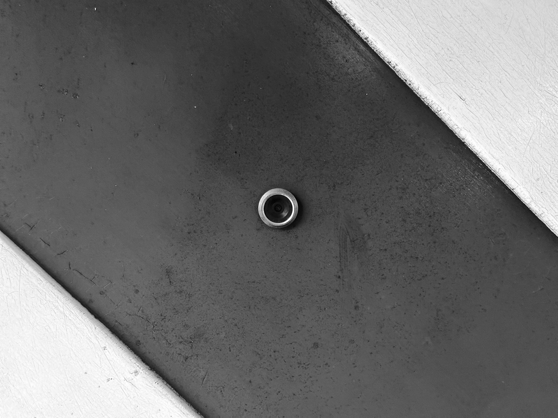 A black and white close up of a security camera lens peaking through an old wooden door, decorated by diagonal light wood panels, the grime of years surrounds the lens, the wood and paint cracks and peels.