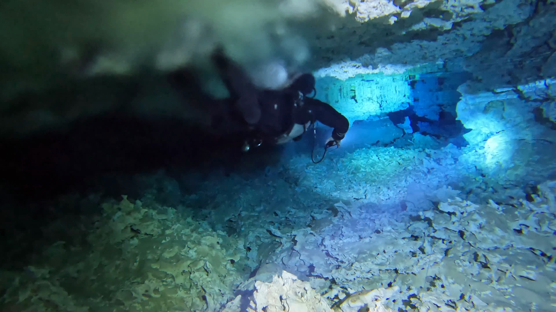 A diver swimming through a brightly lit white pock marked cave, through the middle of the halocline mixing the fresh and salt water layers, trailing a blurry interface behind them.