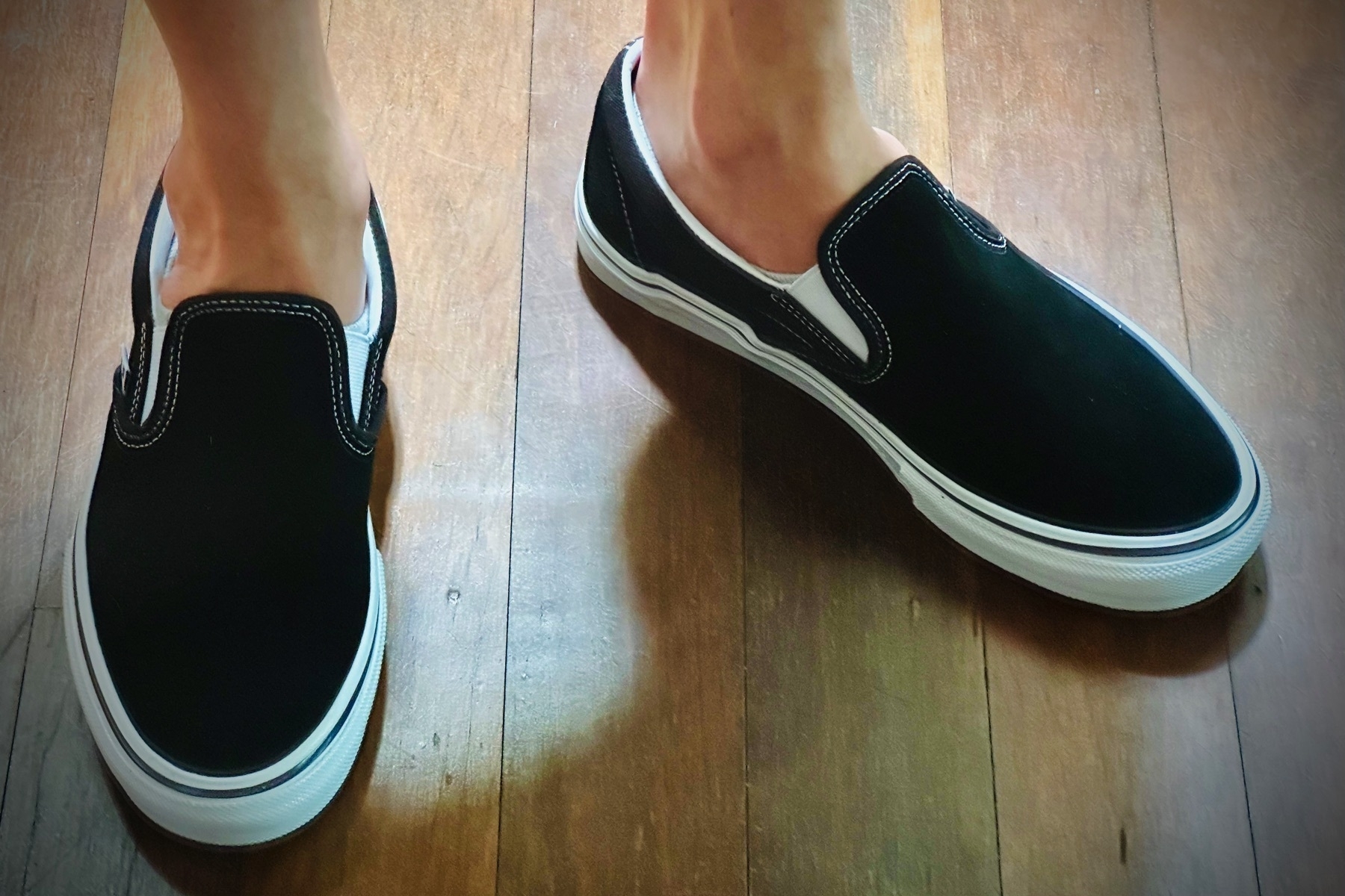 Close of ankles and feet in Vans Skate Sip-on Pro kicks, invisible socks, on a dark polished wood floor.