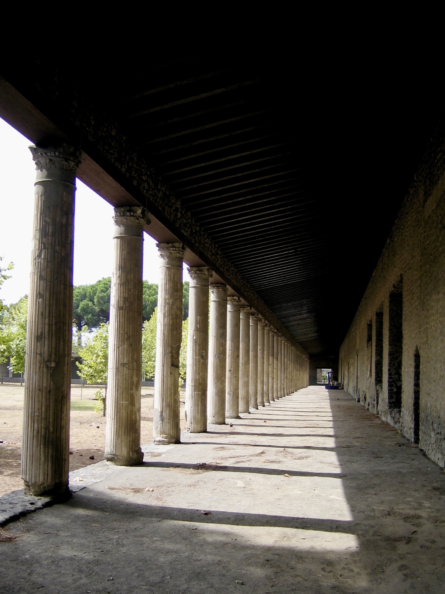 A long portico framed by stone columns stretches into the distance, sunlight streaming through the columns creating stark shadows down its length.