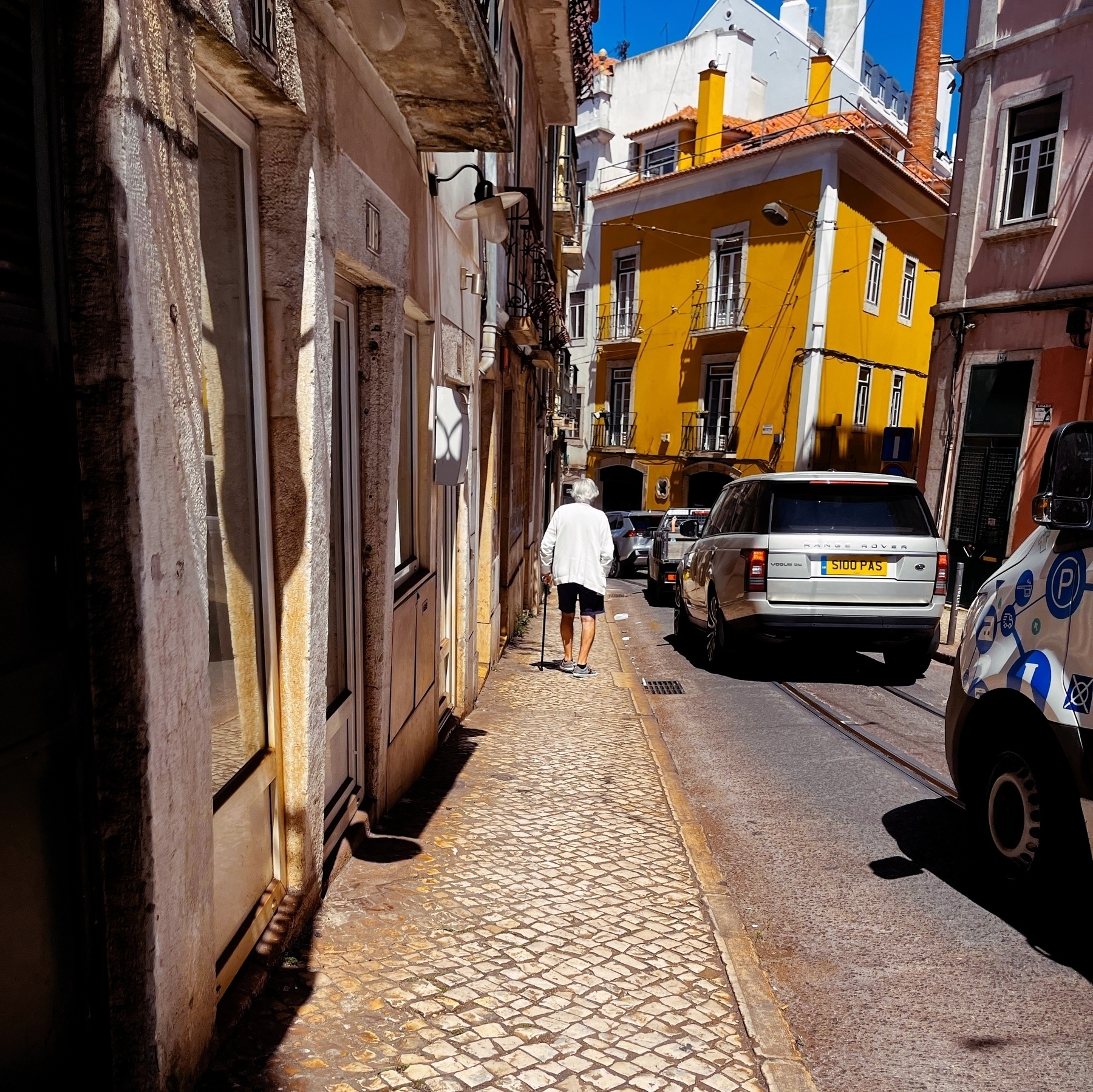 Old man wearing a white cardigan and black shorts, out for a walk in the narrow streets of Lisbon, steadying himself with a cane. Stationary traffic clogs the single lane road. 
