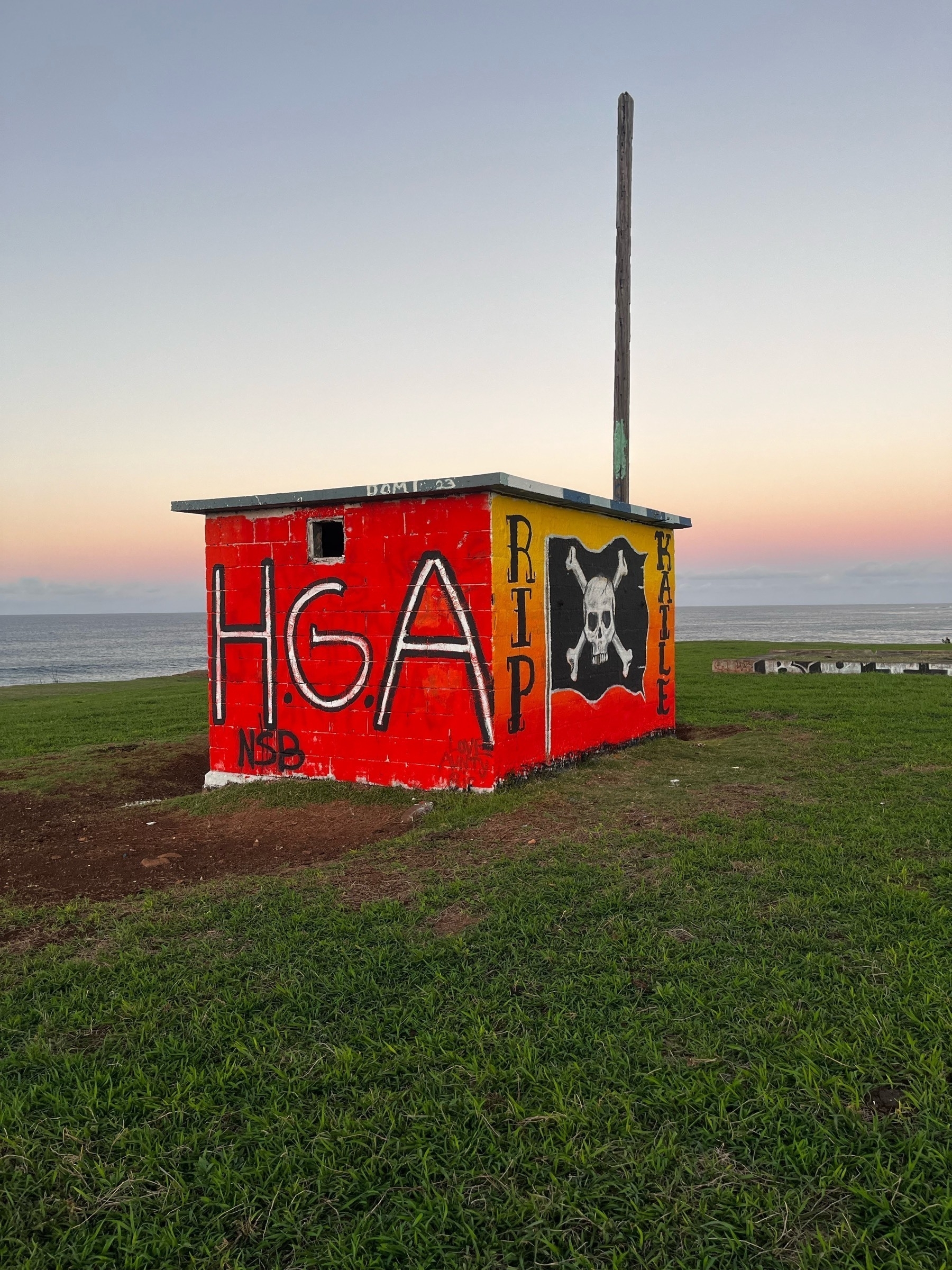 Small red graffiti-covered concrete building with a tall pole on top, set against a coastal landscape with grass in the foreground and a sunset sky in the background.