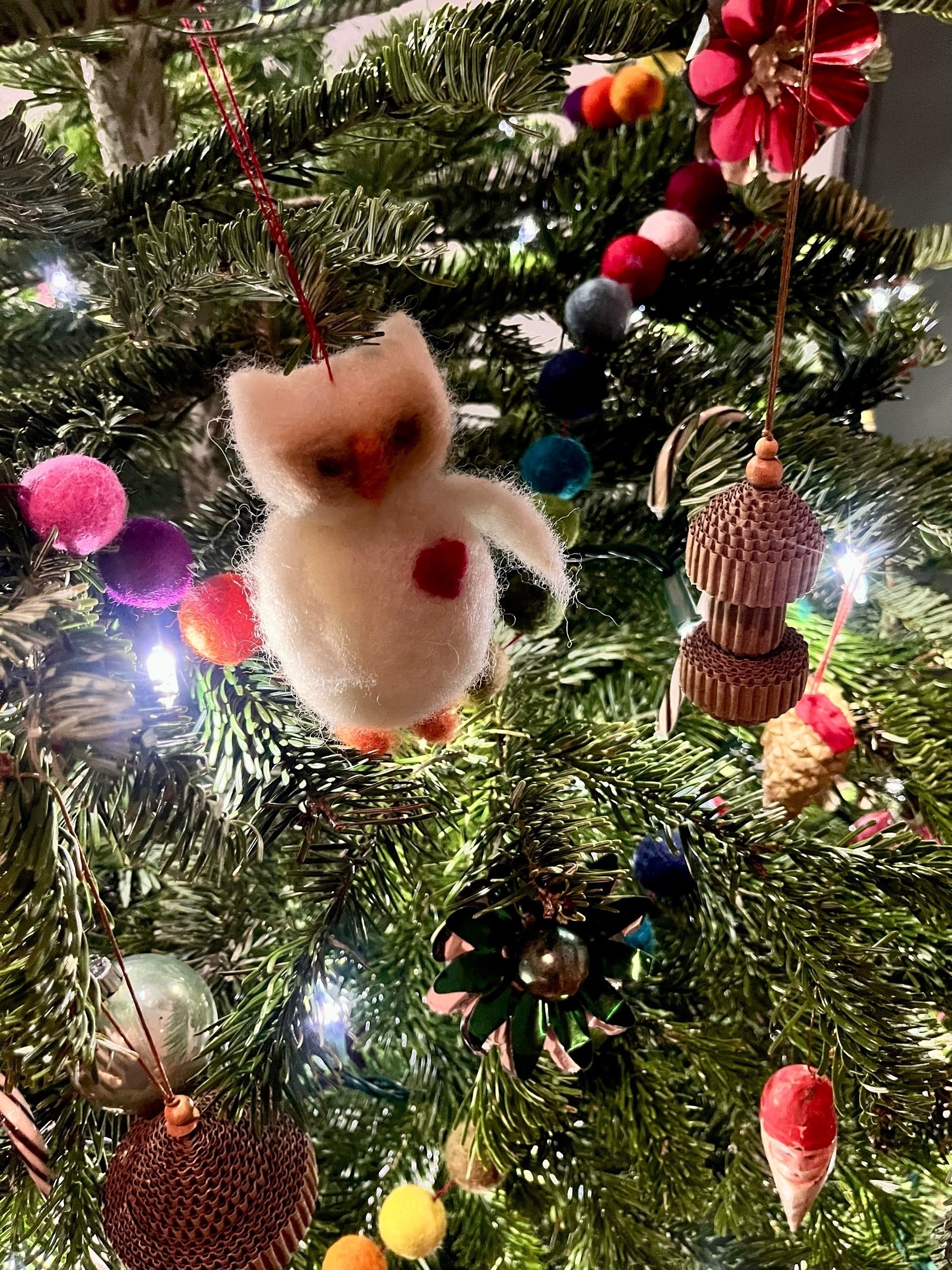 Close-up of a Christmas tree with various decorations including a felted owl ornament, a string of colorful felt balls, and miniature Santa Clauses. 
