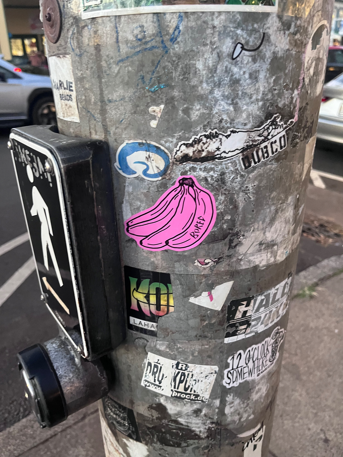 Stickers, including some pink bananas, on a pole at a street light crossing
