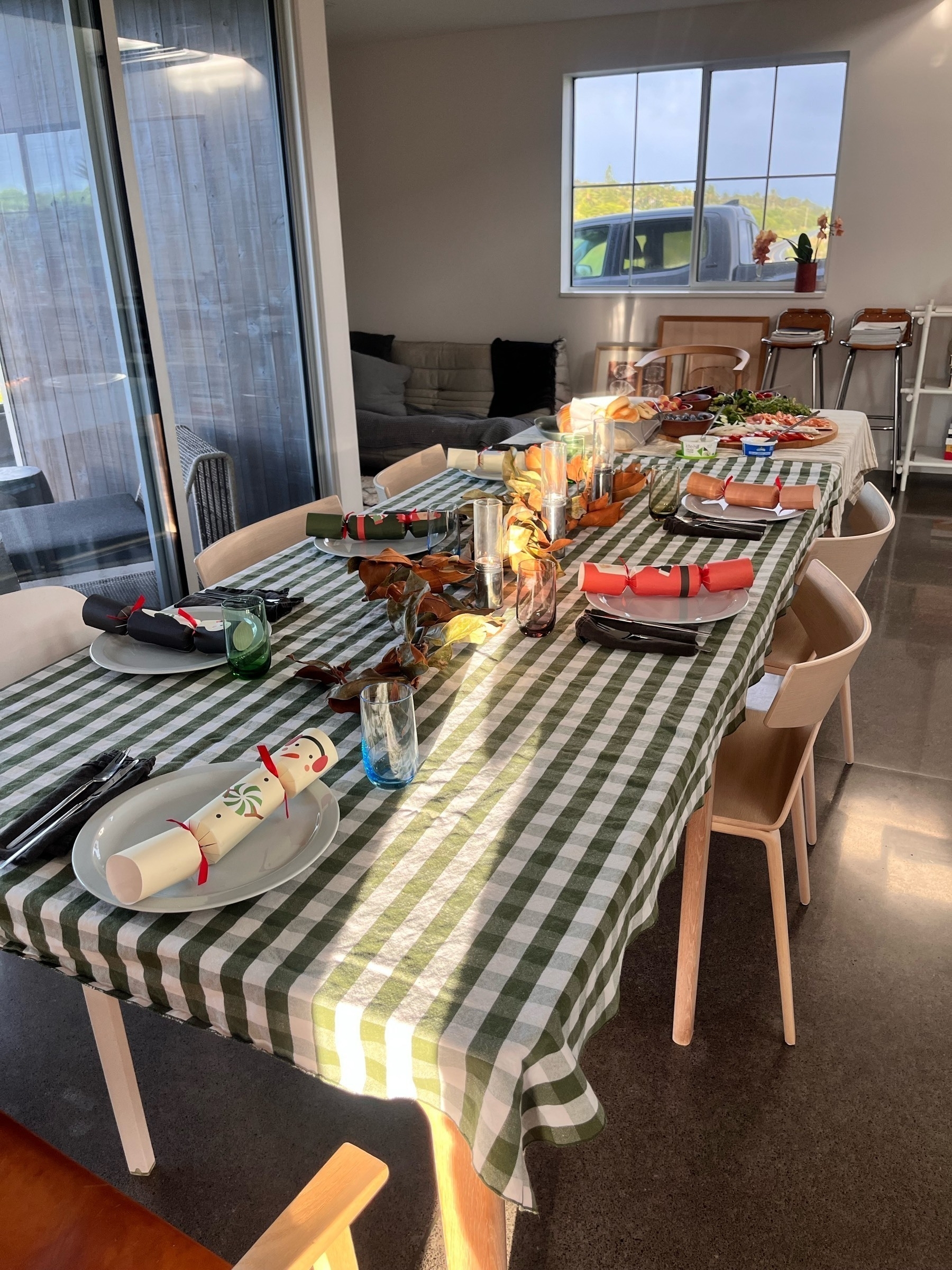 A table laid out for breakfast. In each plate is a Christmas cracker. At the far end of the table is the food, so people can help themselves buffet style. Sun light shines the length of the table