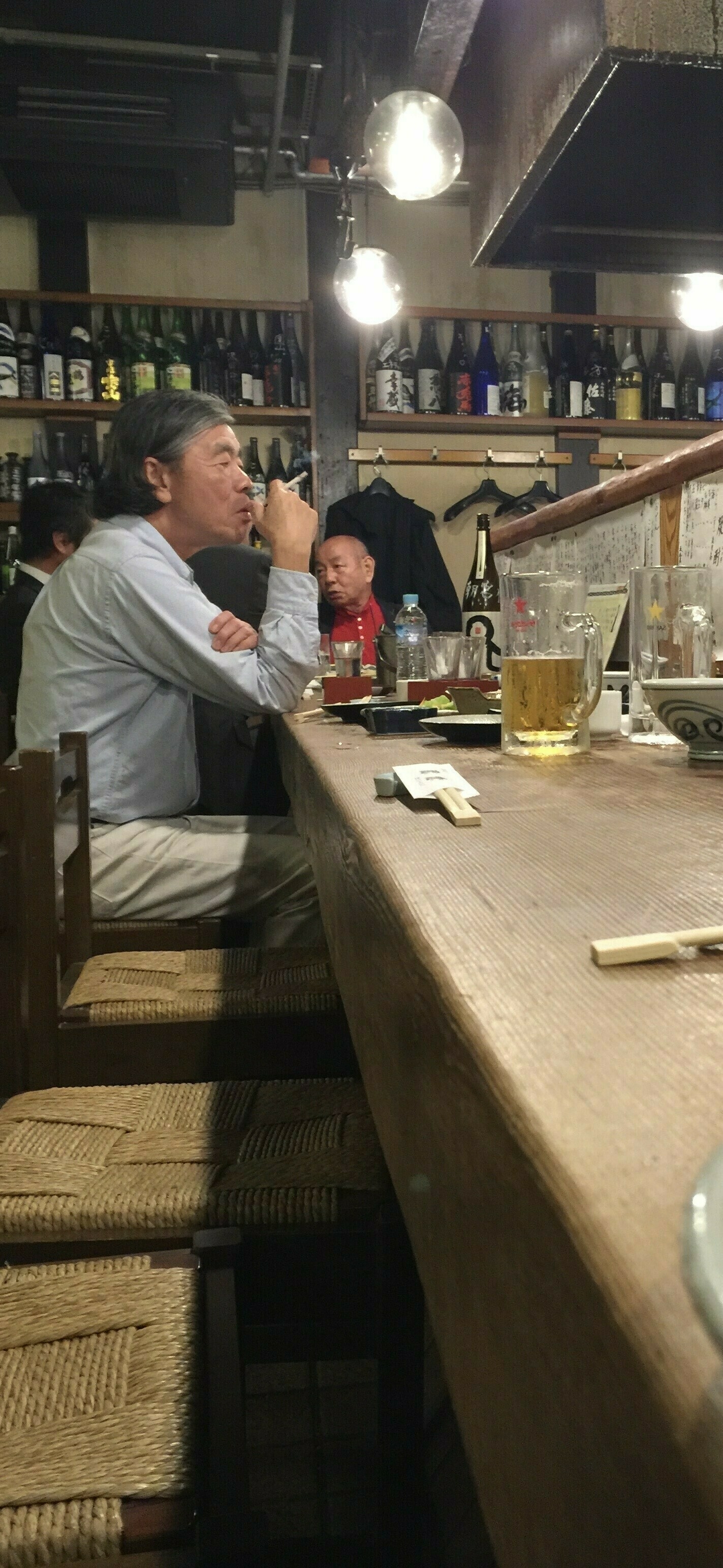 Japanese men sitting at a bar in Japan. One is smoking. A half drunk glass of beer sits nearby
