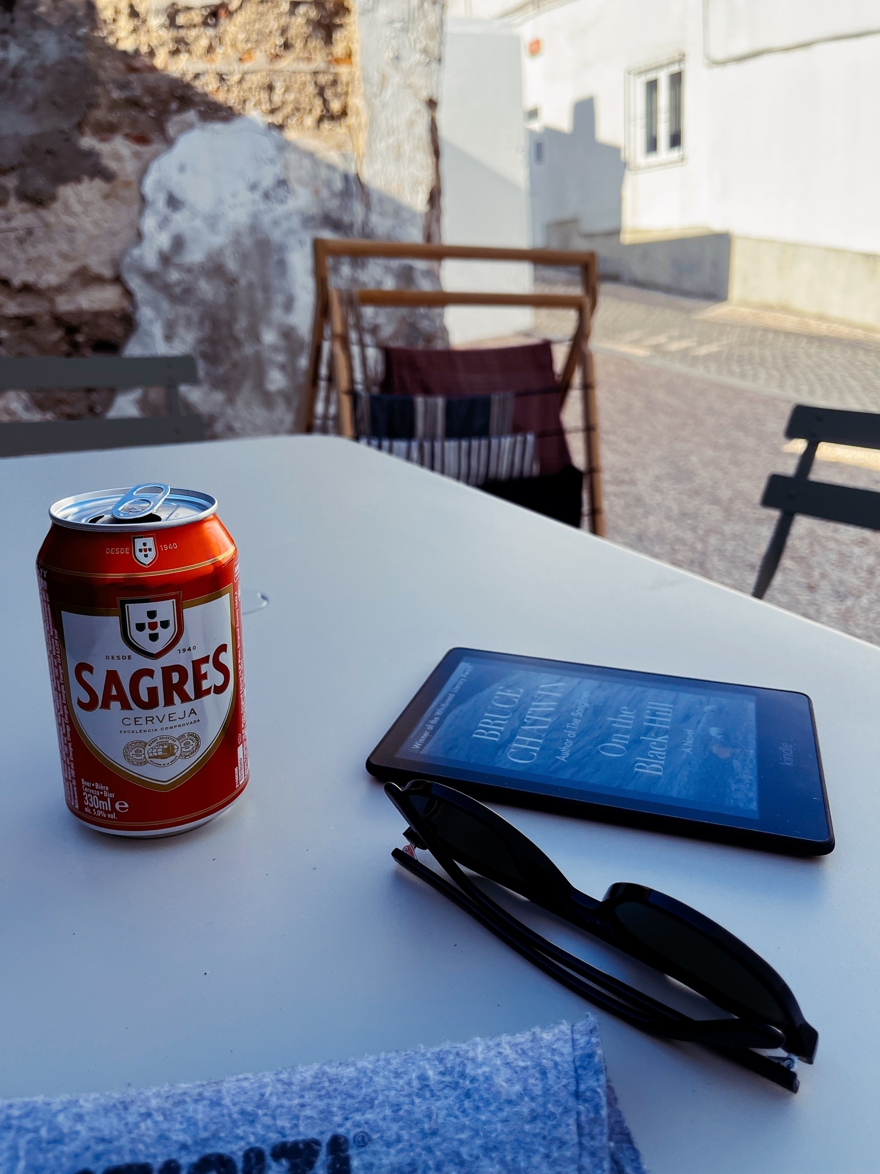 Sitting outside next to a table with my Kindle in it, along with sun glasses, a can of beer, and a soft glasses case. Laundry is hanging on a clothes horse, just out of focus. 