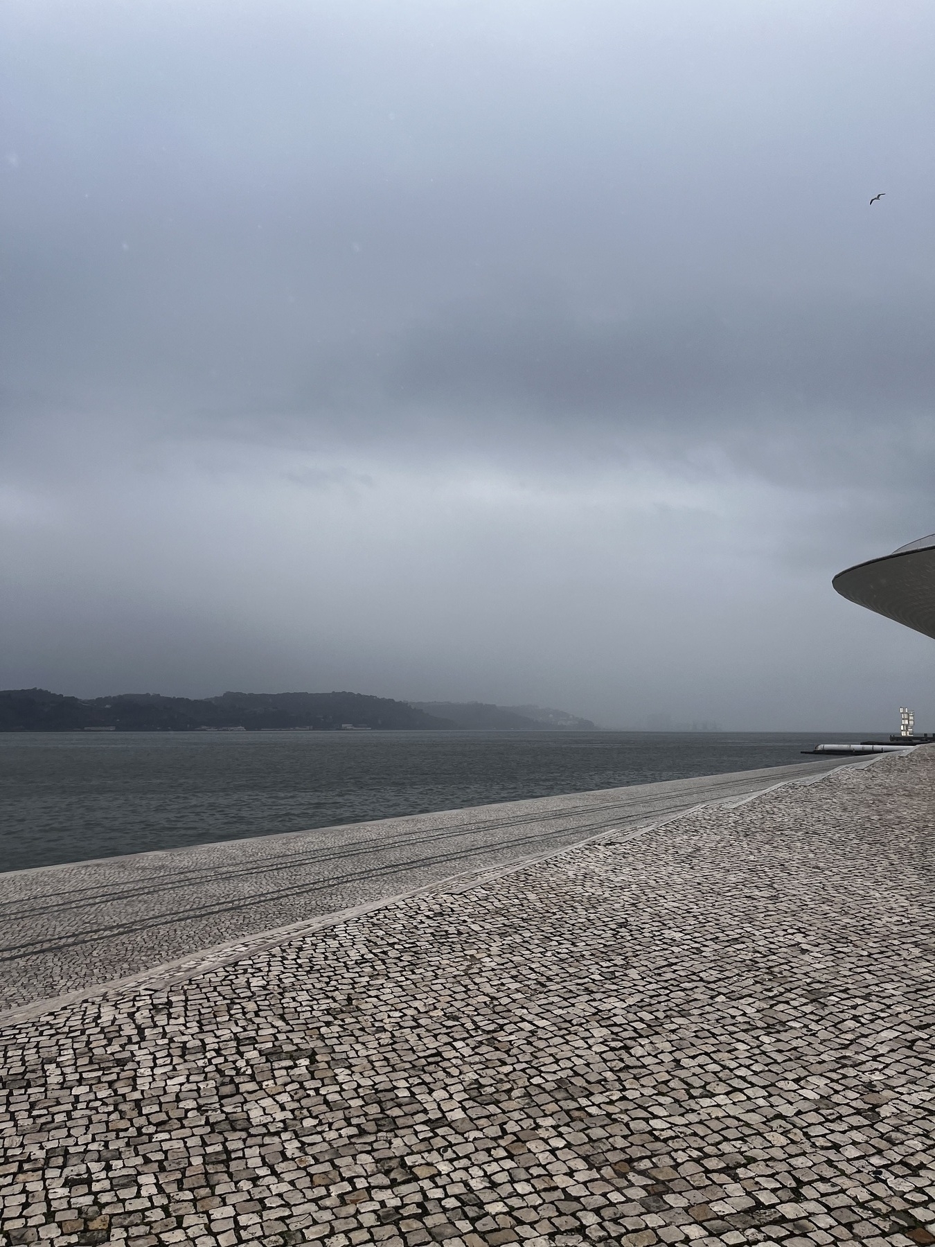 A cloudy, overcast sky looms over a cobblestone walkway beside a calm body of water.