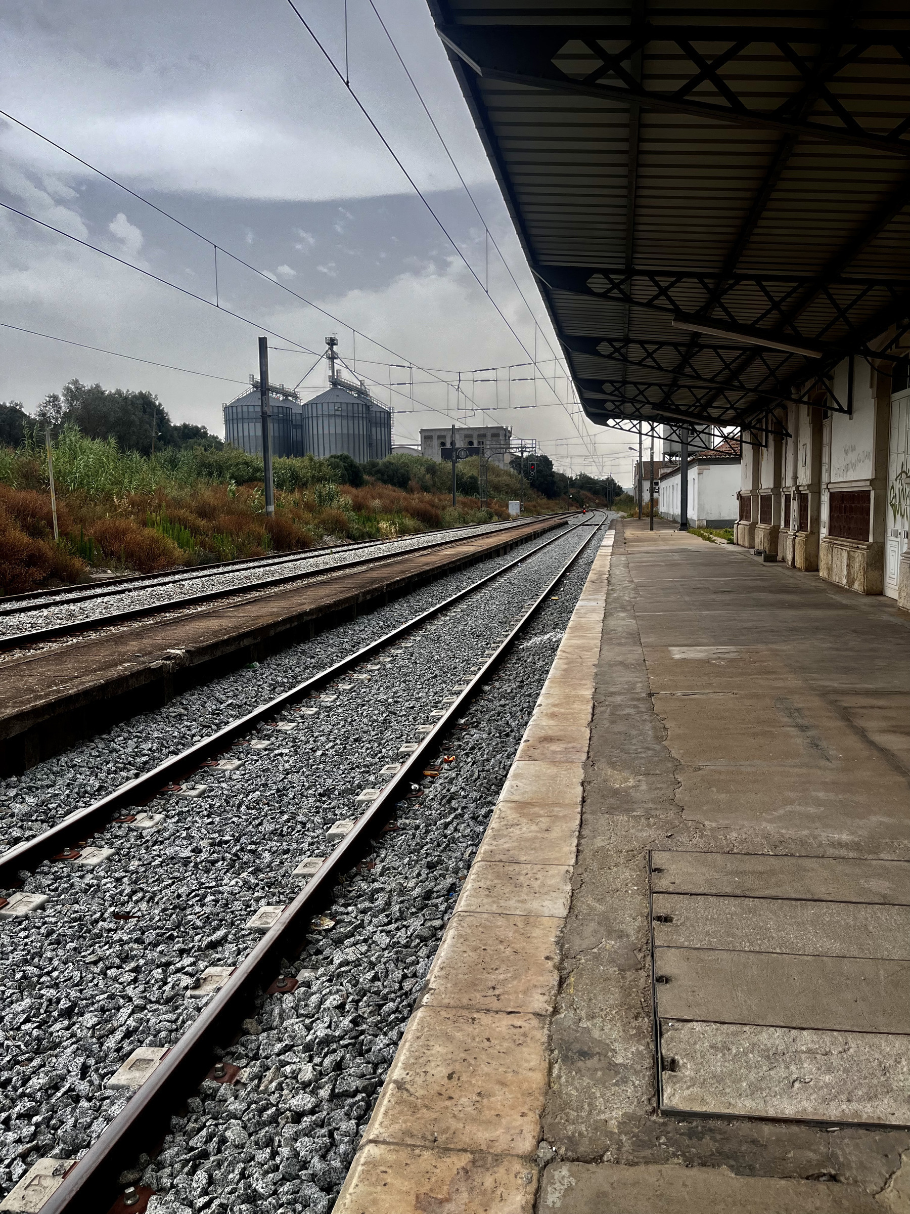 Taken in a railway station. Train tracks disappear into the distance from a covered platform. On the far side is a grass bank. In the distance some silos, maybe rice silos.