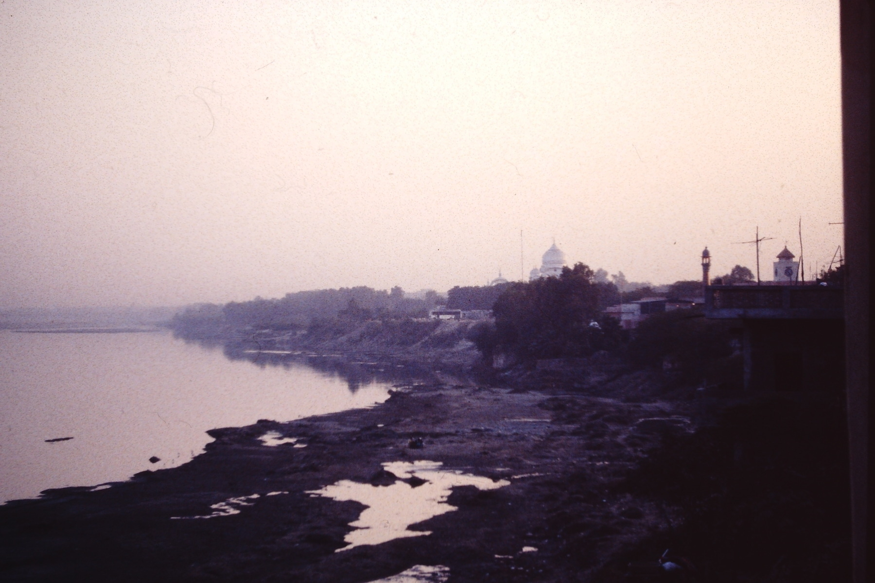 A calm river bends around a hazy landscape with buildings in the distance and a rocky shoreline in the foreground.