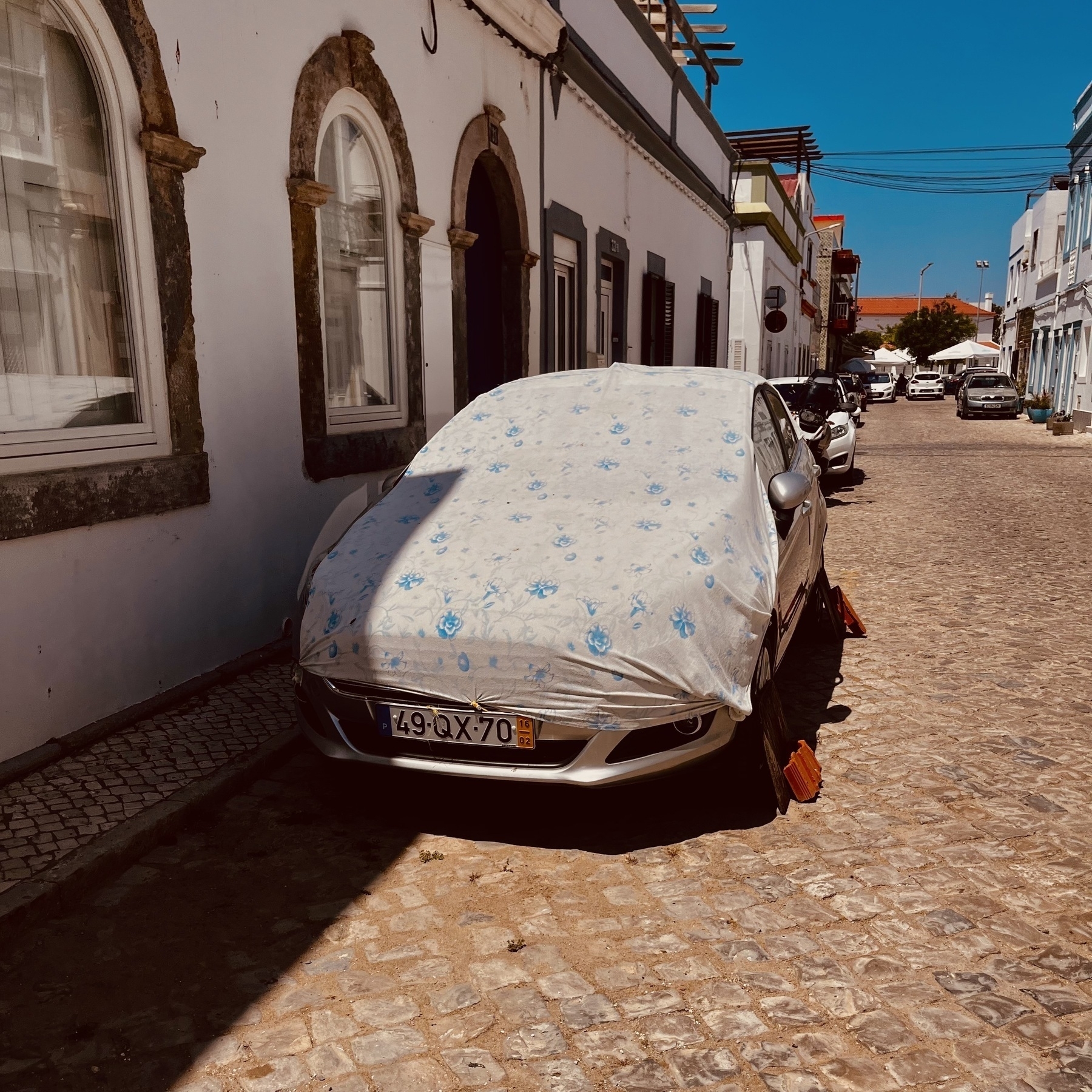 A car parked on a cobbled street with terraced houses on both sides. The car is covered in a sheet to protect it from the heat of the sun.