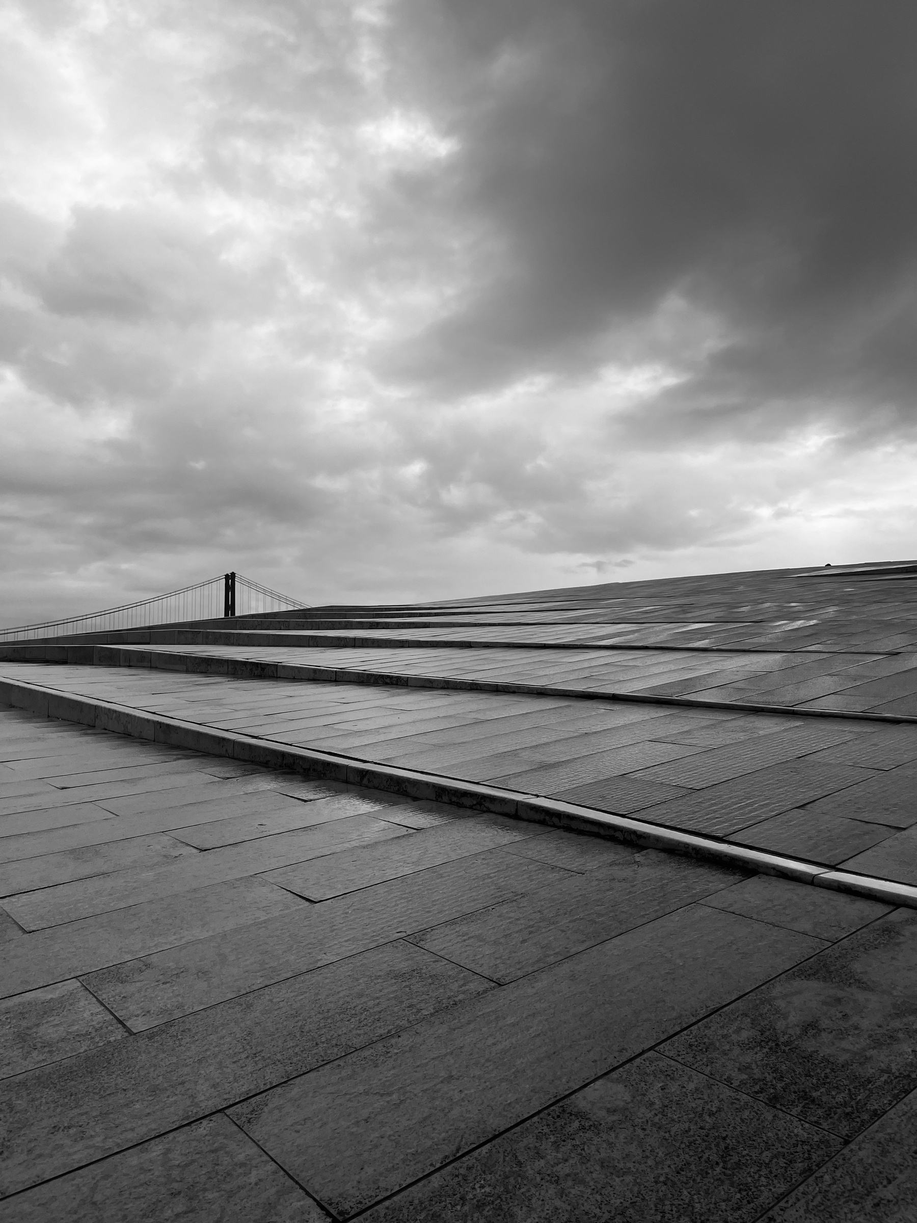 A dramatic black-and-white scene features a cloudy sky above a modern, angular building with the outline of a bridge visible in the distance.