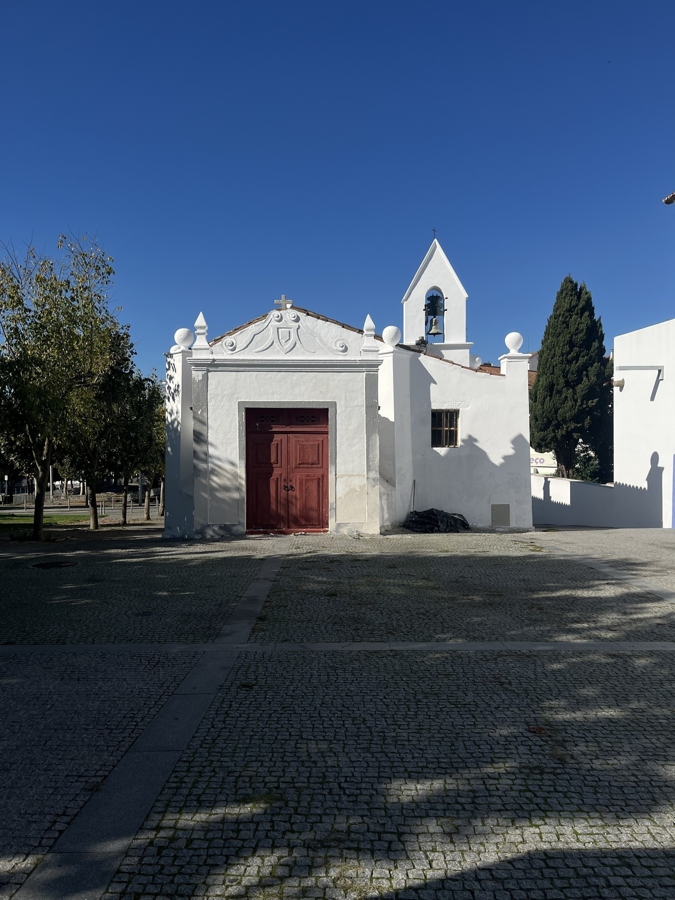 A small, white chapel with a red door and a bell tower is situated in a sunny cobblestone courtyard surrounded by trees.