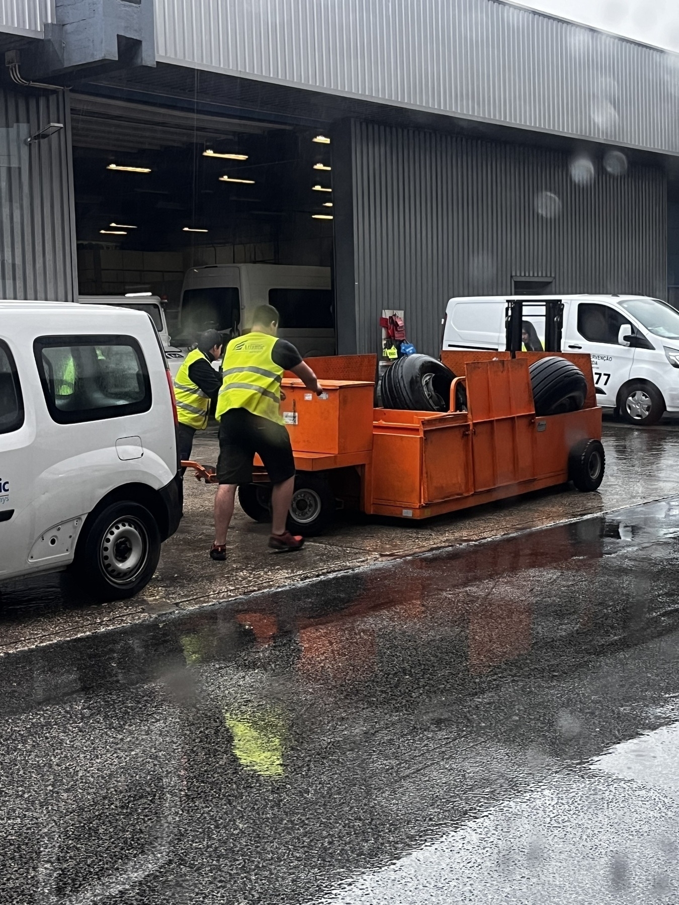 Workers in high-visibility vests are moving stacks of tires using an orange cart outside a garage.