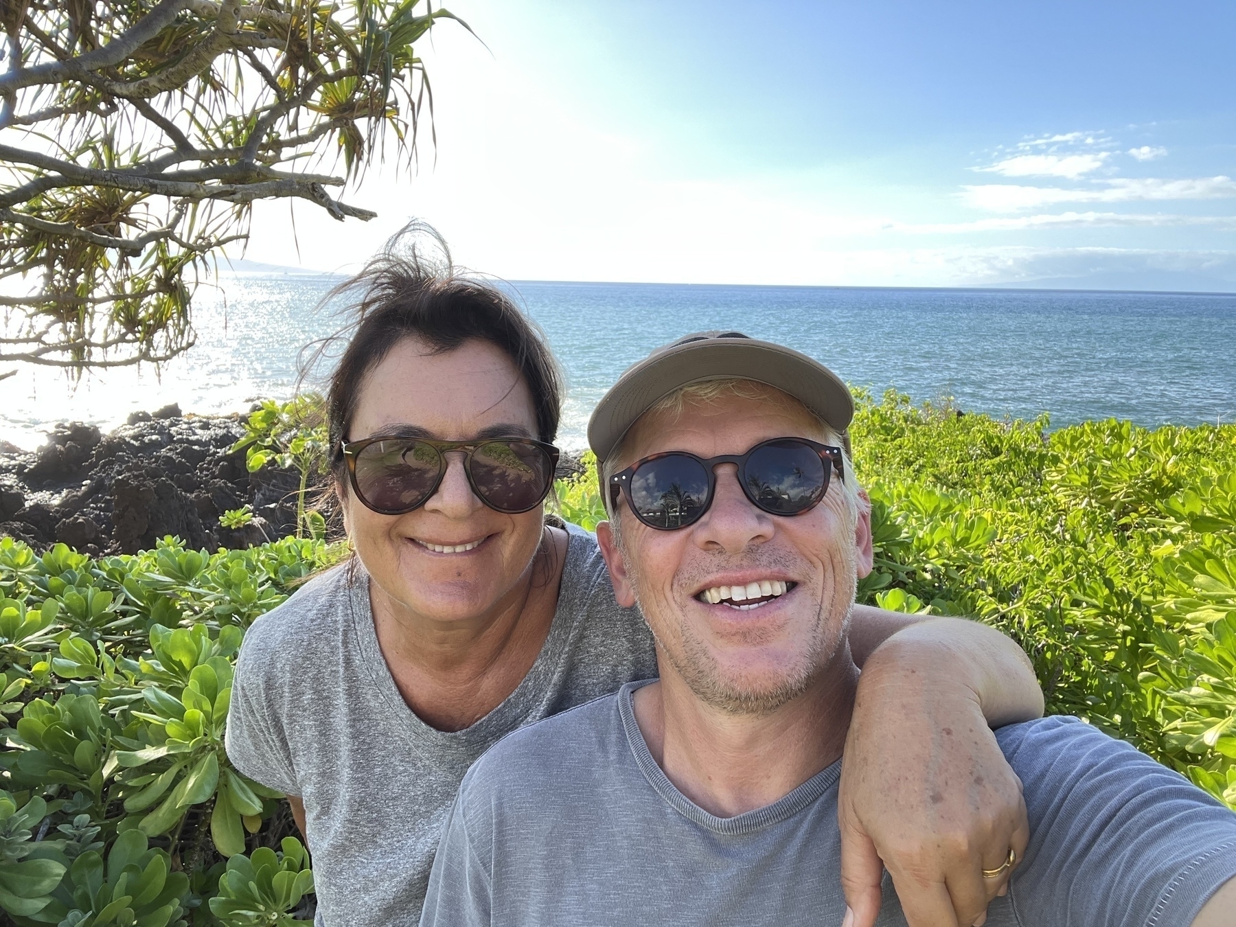 A couple wearing sunglasses smiles at the camera in a lush coastal landscape with the ocean in the background.