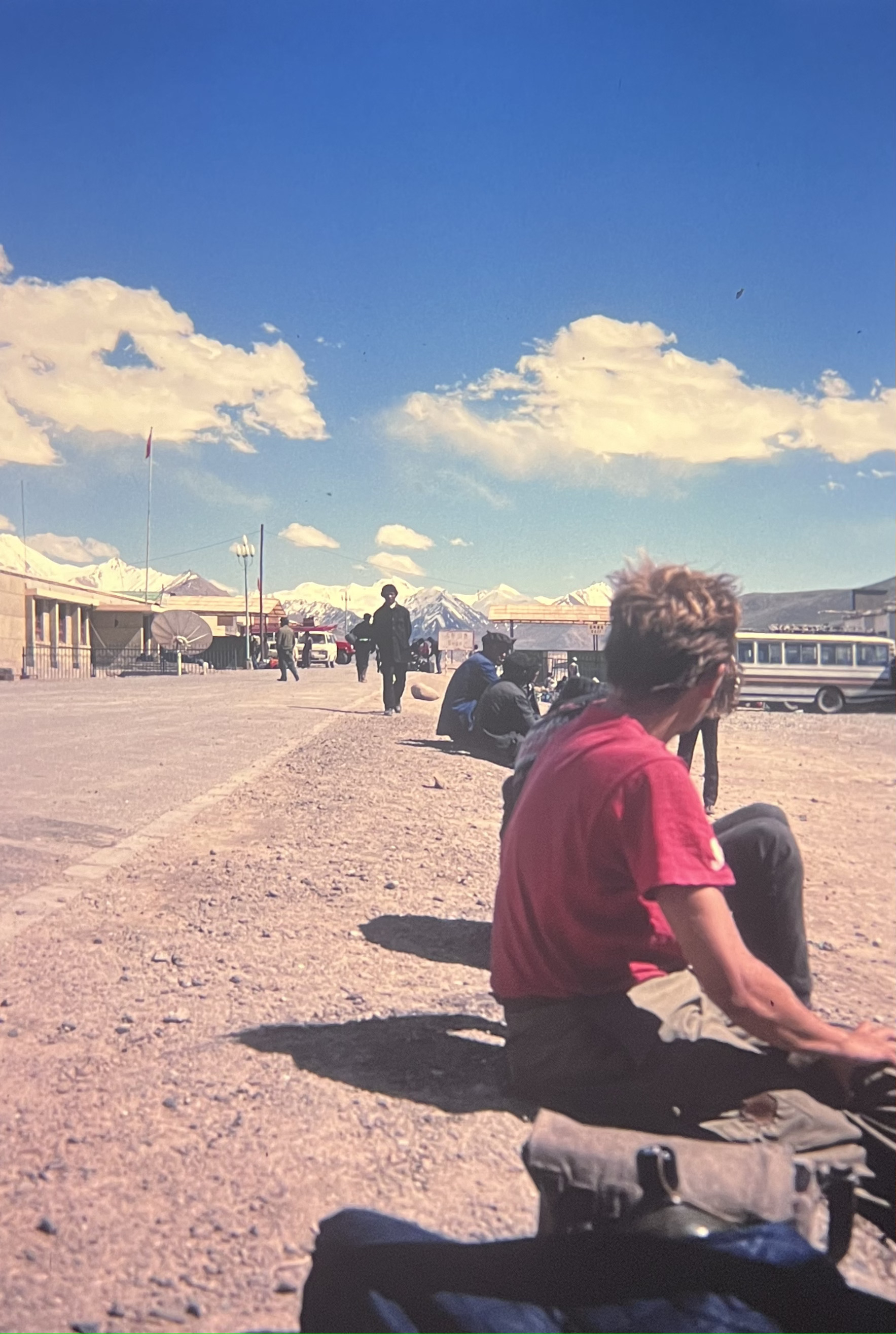 A person in a red shirt sits on the ground near a road with several others in the distance, under a bright blue sky with scattered clouds.