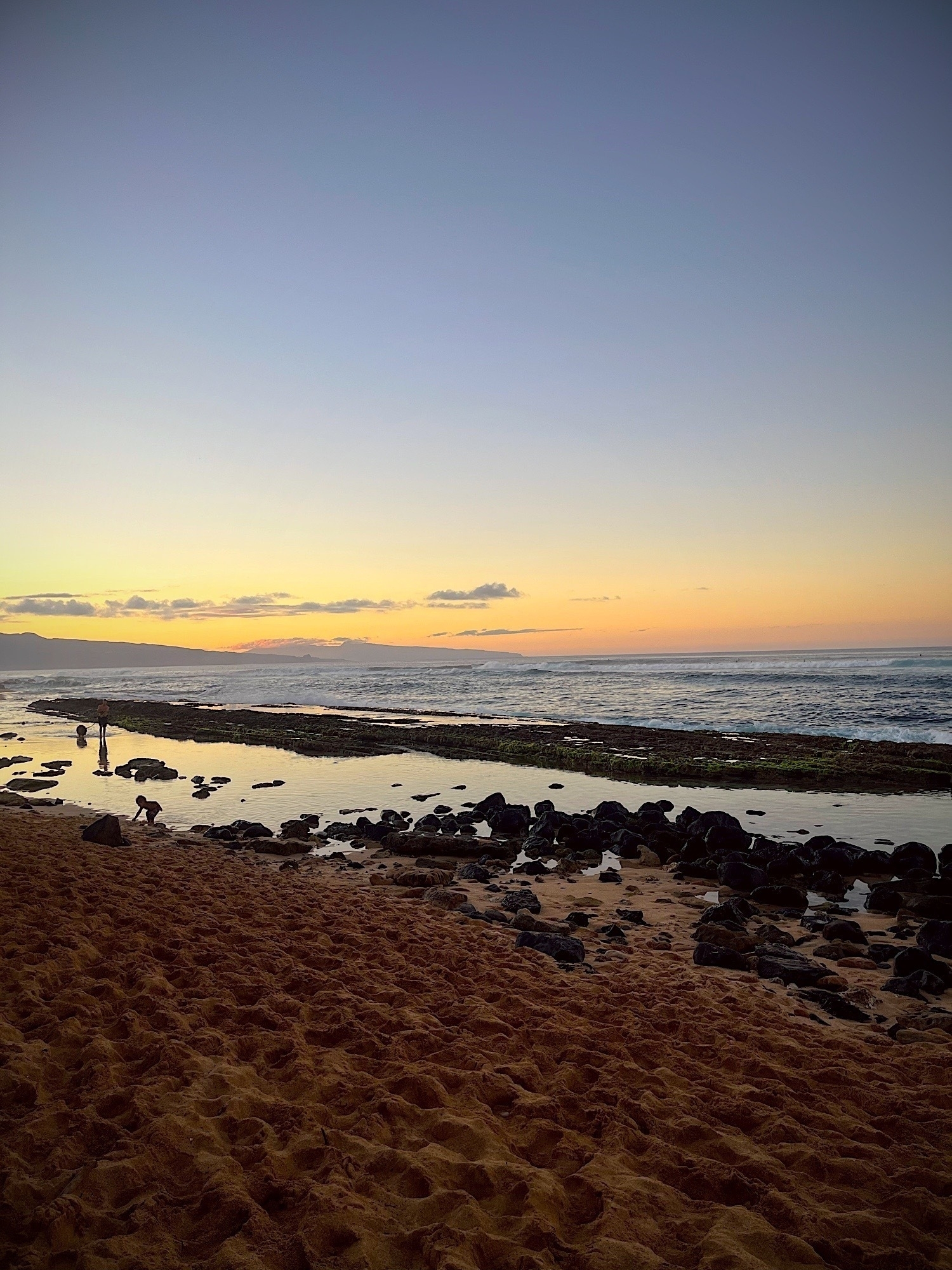 The orange and yellow glow of sunset over the ocean. A cloudless sky, rocks and pools in the middle ground with a beach in the foreground.