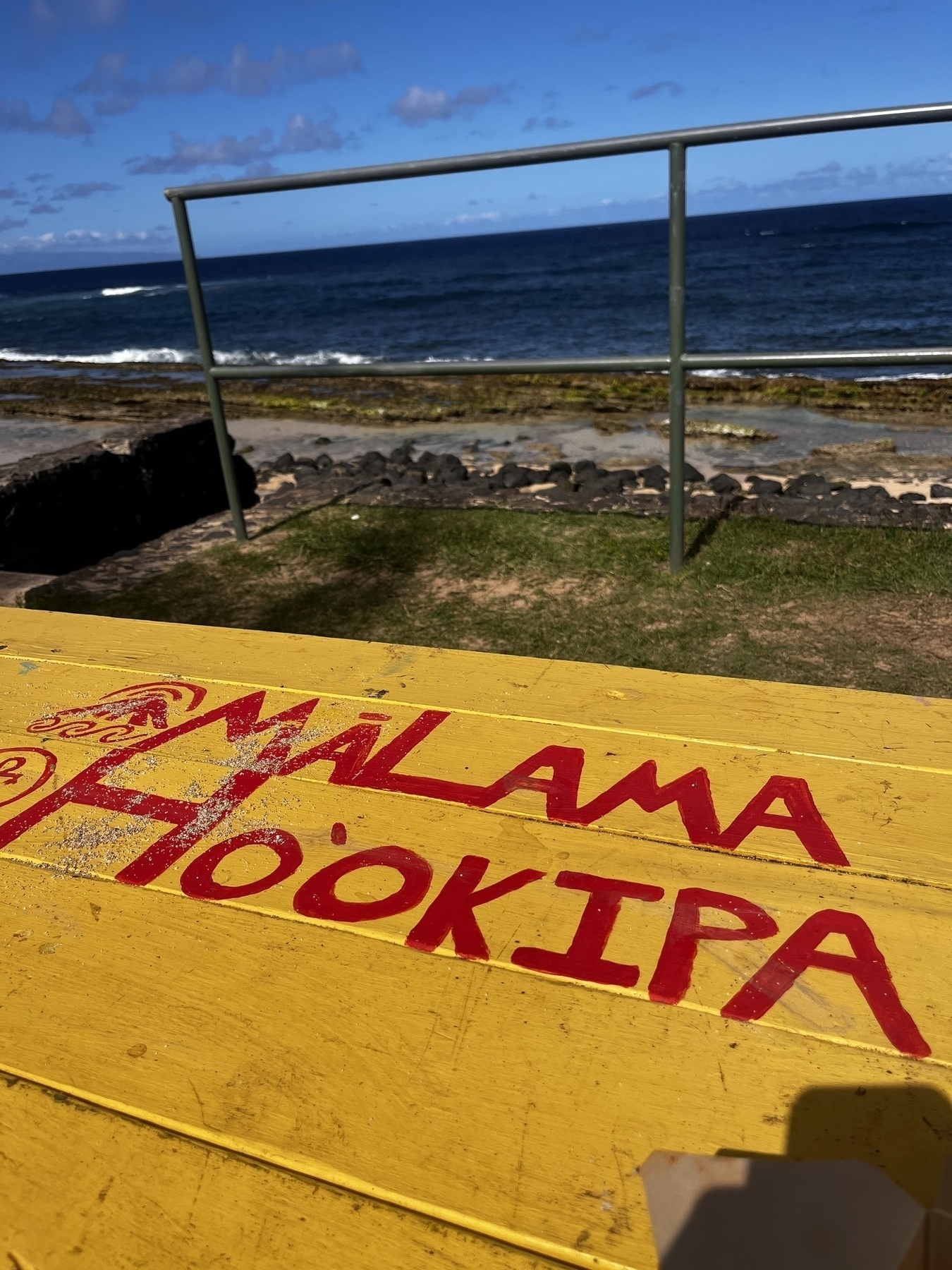 A yellow table with red text saying MALAMA HO’OKIPA overlooks a rocky shoreline and the ocean.