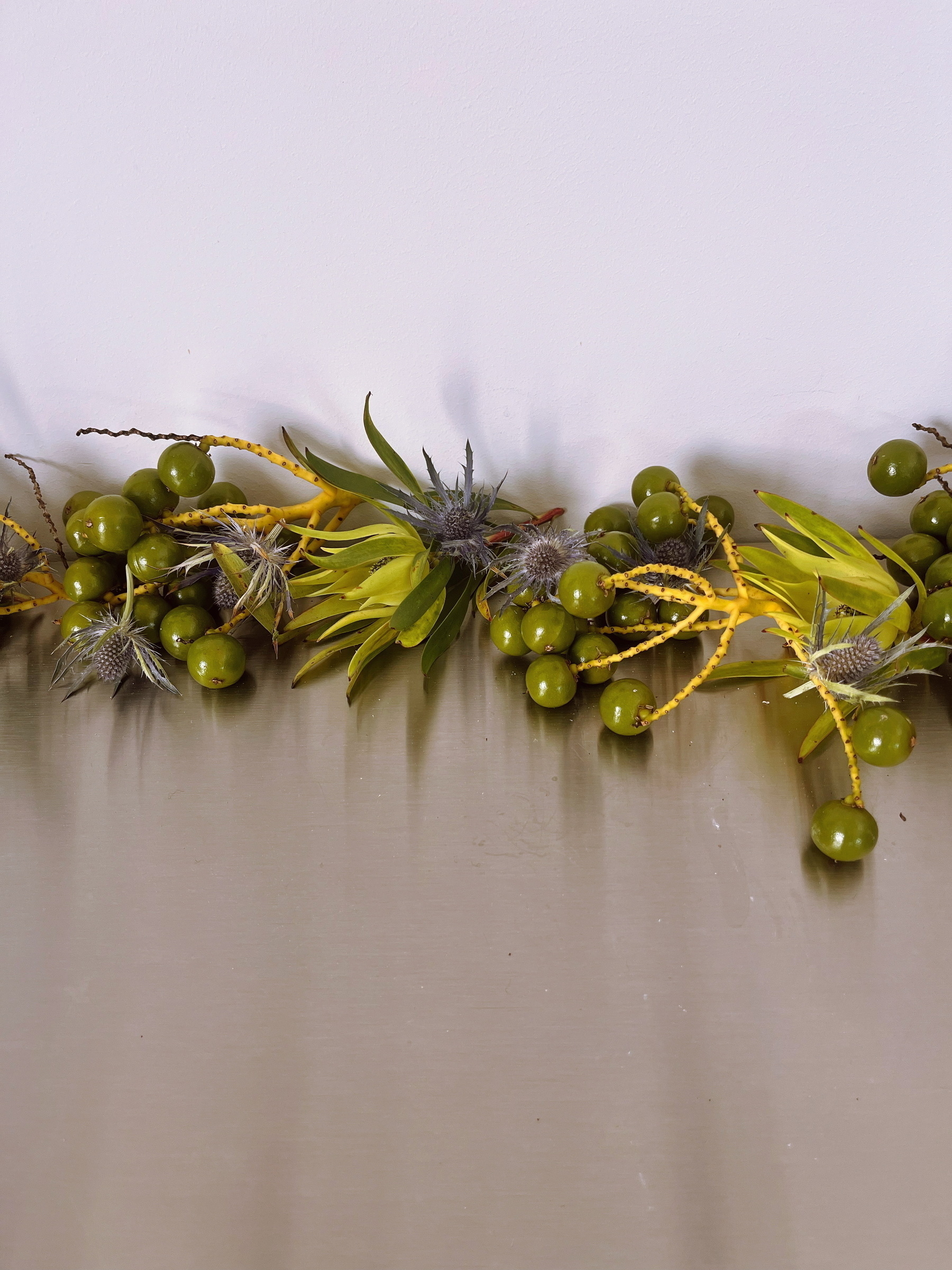 A tabletop arrangement features clusters of green berries, yellow accents, and spiky, thistle-like flowers.