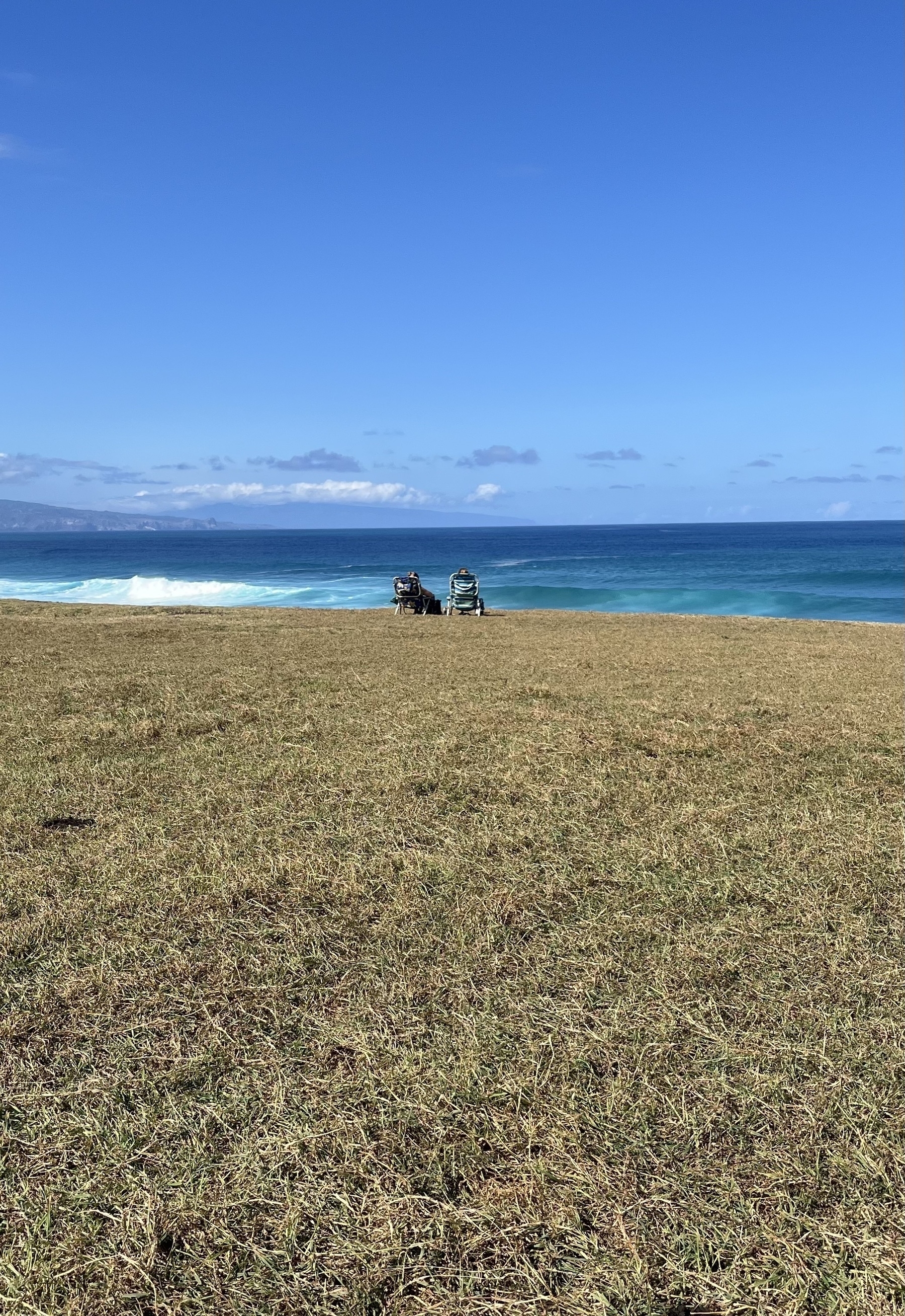 Two people sit in chairs on a grassy hill overlooking a blue ocean and clear sky.