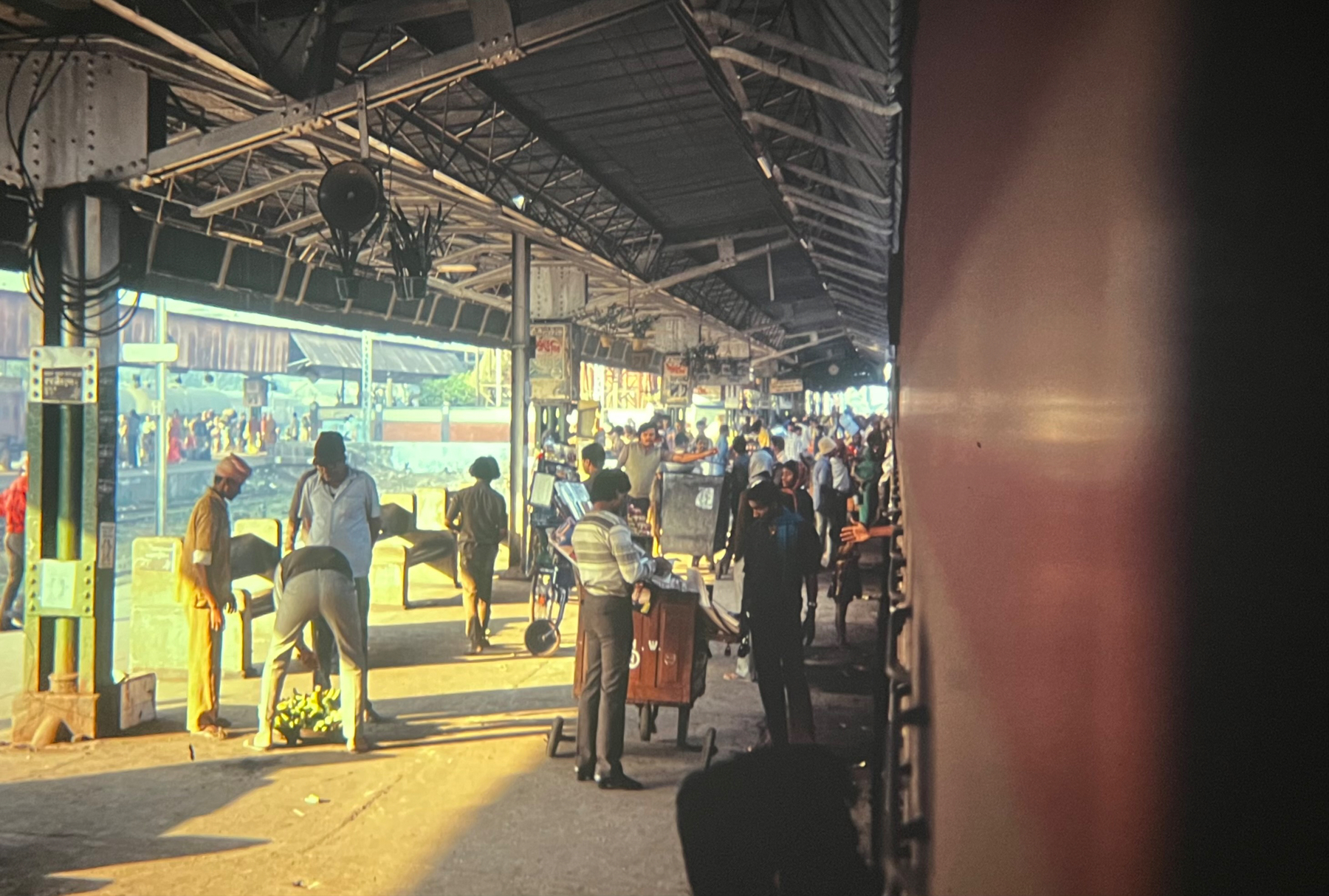 A bustling train station in India with passengers walking, standing, and talking; some are seated waiting for trains. Platform shelters and station signage are visible, alongside luggage and a vendor cart. The view is framed by a train on the right edge.