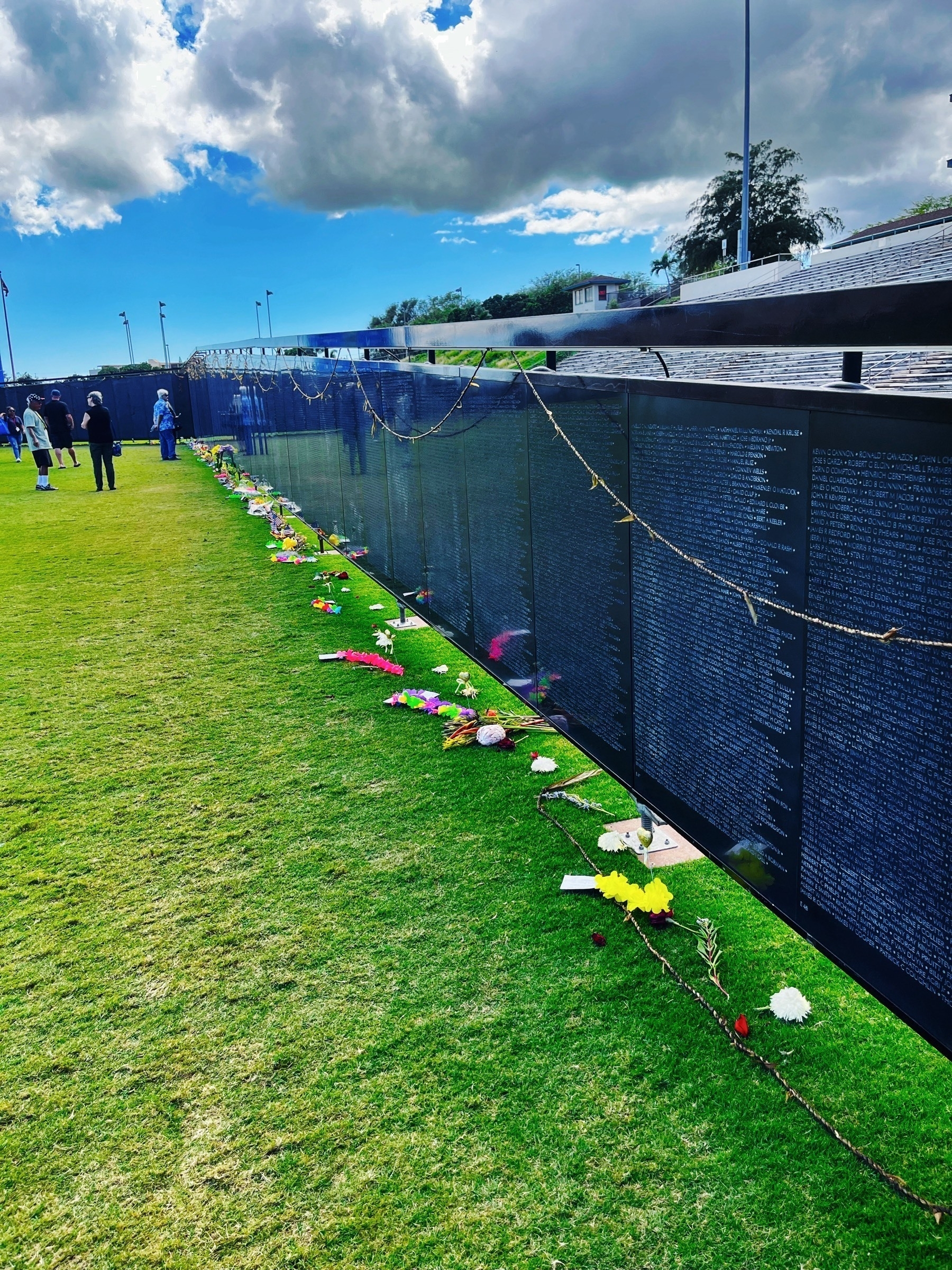 A wall standing on grass with names on it extends to the distance. Leis are draped over it, flower offerings on the grass and people in the background