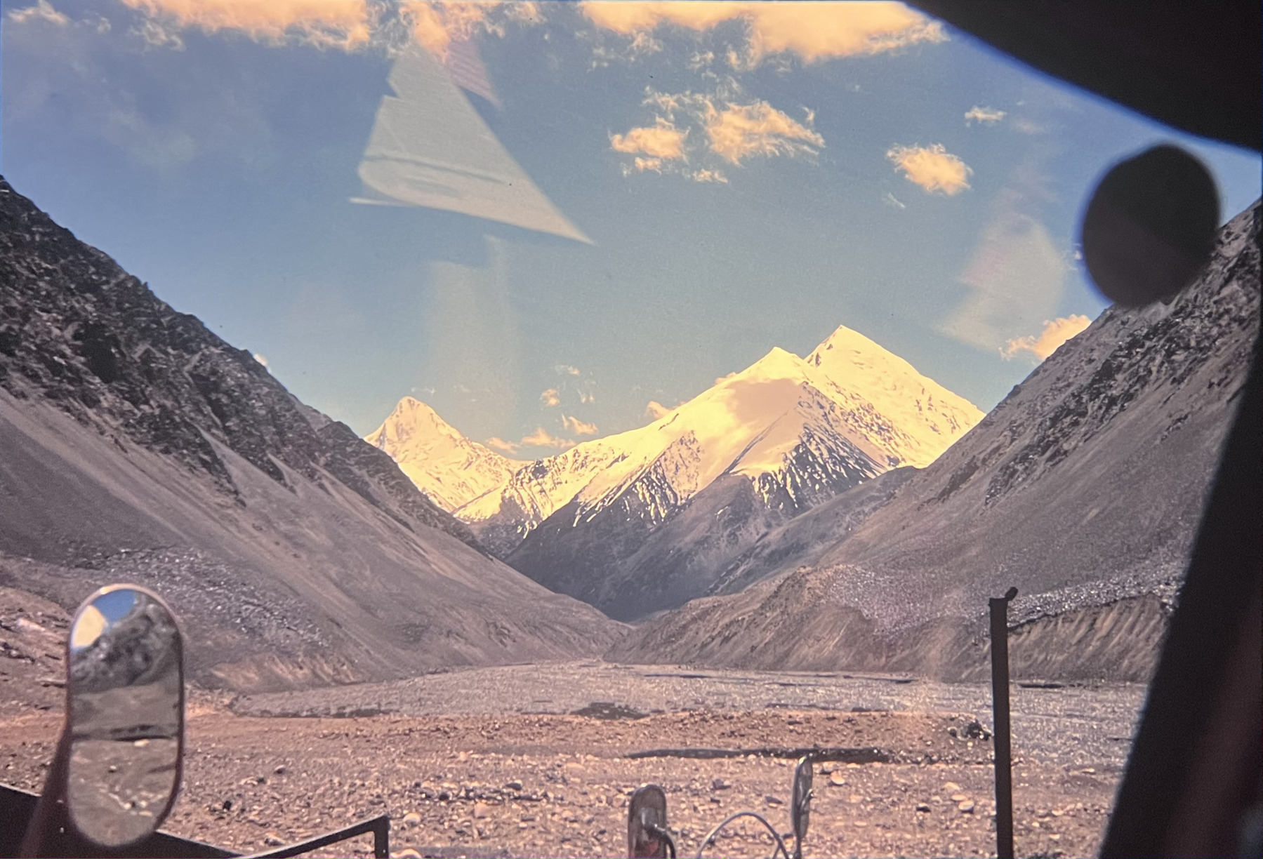 A snow-capped mountain range is visible through the windshield of a vehicle, framed by rugged, rocky terrain.