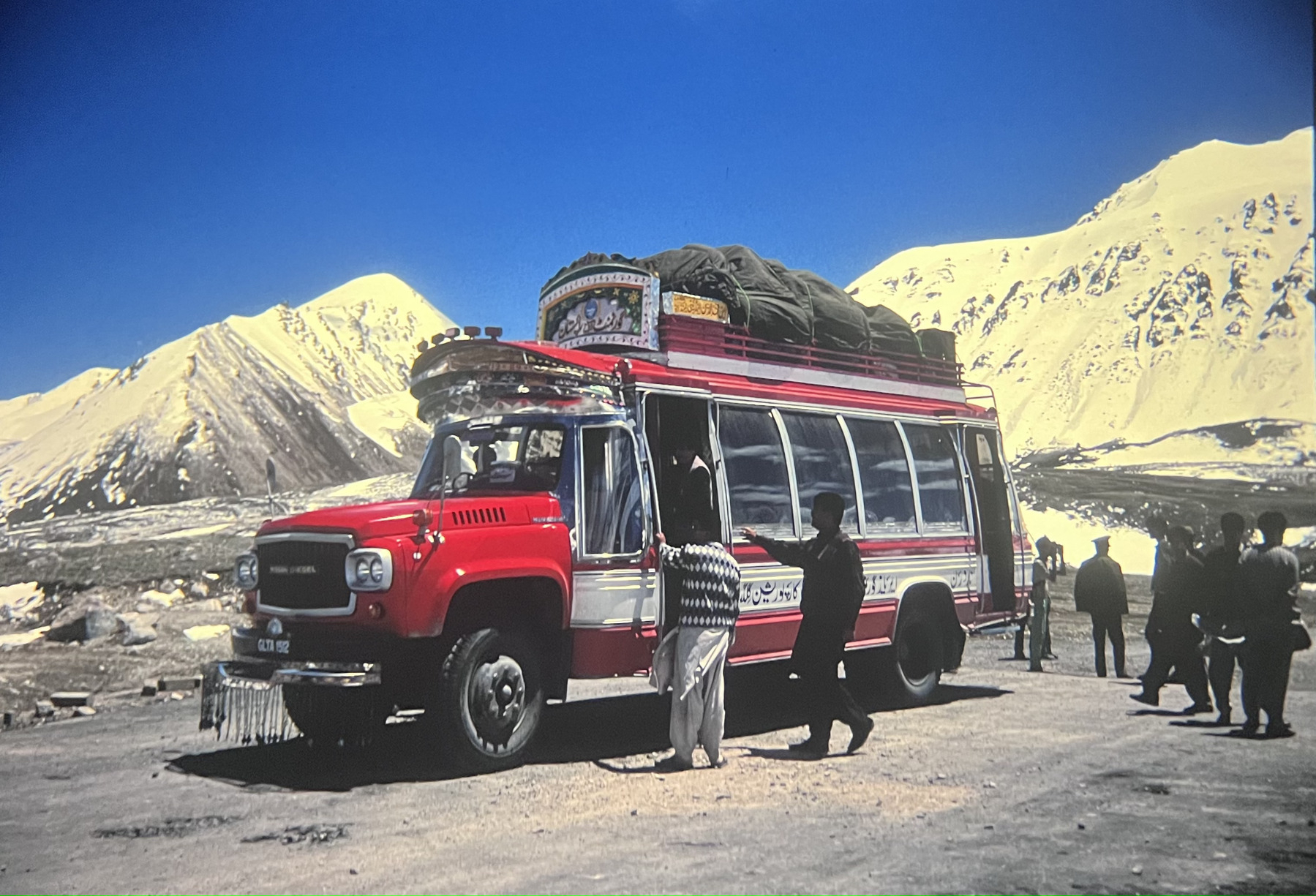 A colourful red and white Pakistan bus parked at the Khunjerab Pass. People walk around with snow covered mountains around